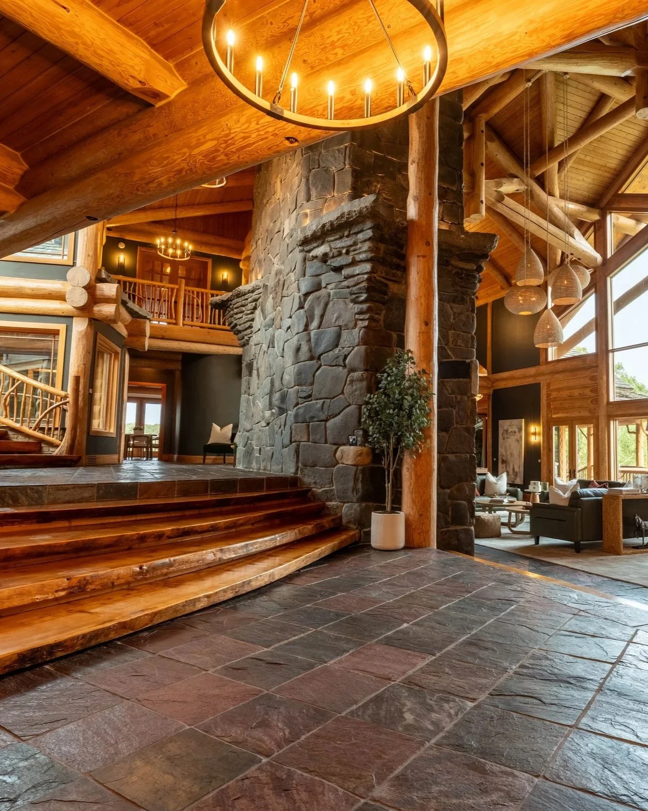 Interior of a rustic lodge or cabin with a large stone fireplace, wooden beams, and high ceilings with multiple windows. There are chandeliers and modern furniture visible in the background.