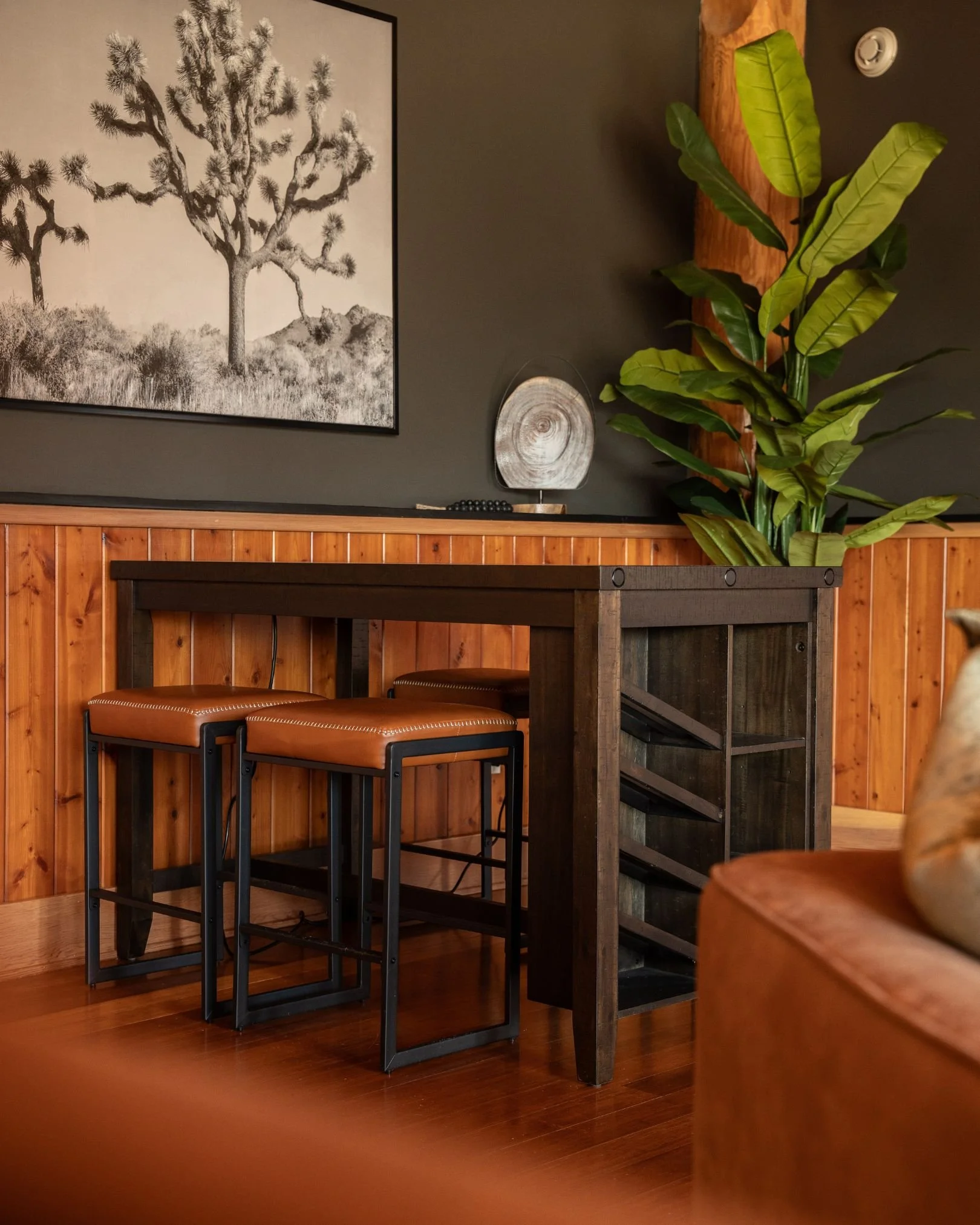 A cozy corner of a room with a black wooden table, brown cushioned stools, a large green leafy plant, a wall-mounted decorative piece resembling a tree cross-section, a black and white landscape art, and a dark wall with wood paneling at the bottom.