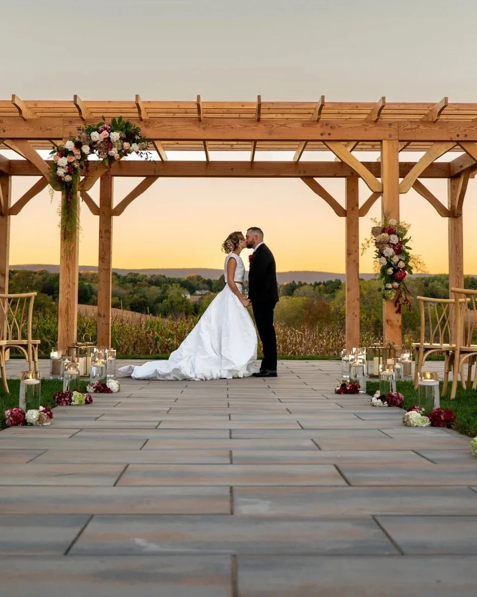 A bride and groom kissing under a pergola during their outdoor wedding at sunset, with flower arrangements and lanterns along the aisle.