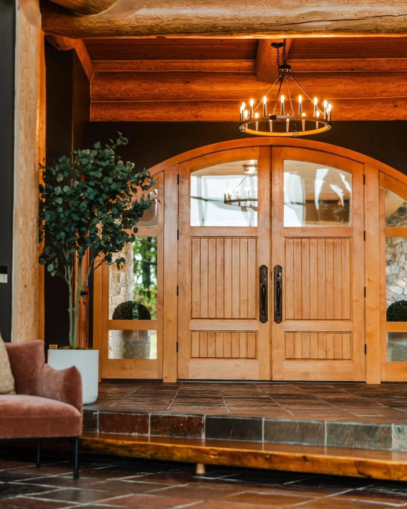 A rustic wooden front door with large glass panes, surrounded by a wooden frame, and a chandelier with candles hanging from the ceiling. A potted plant with green foliage stands near the door, and a brown upholstered chair is partially visible in the foreground.