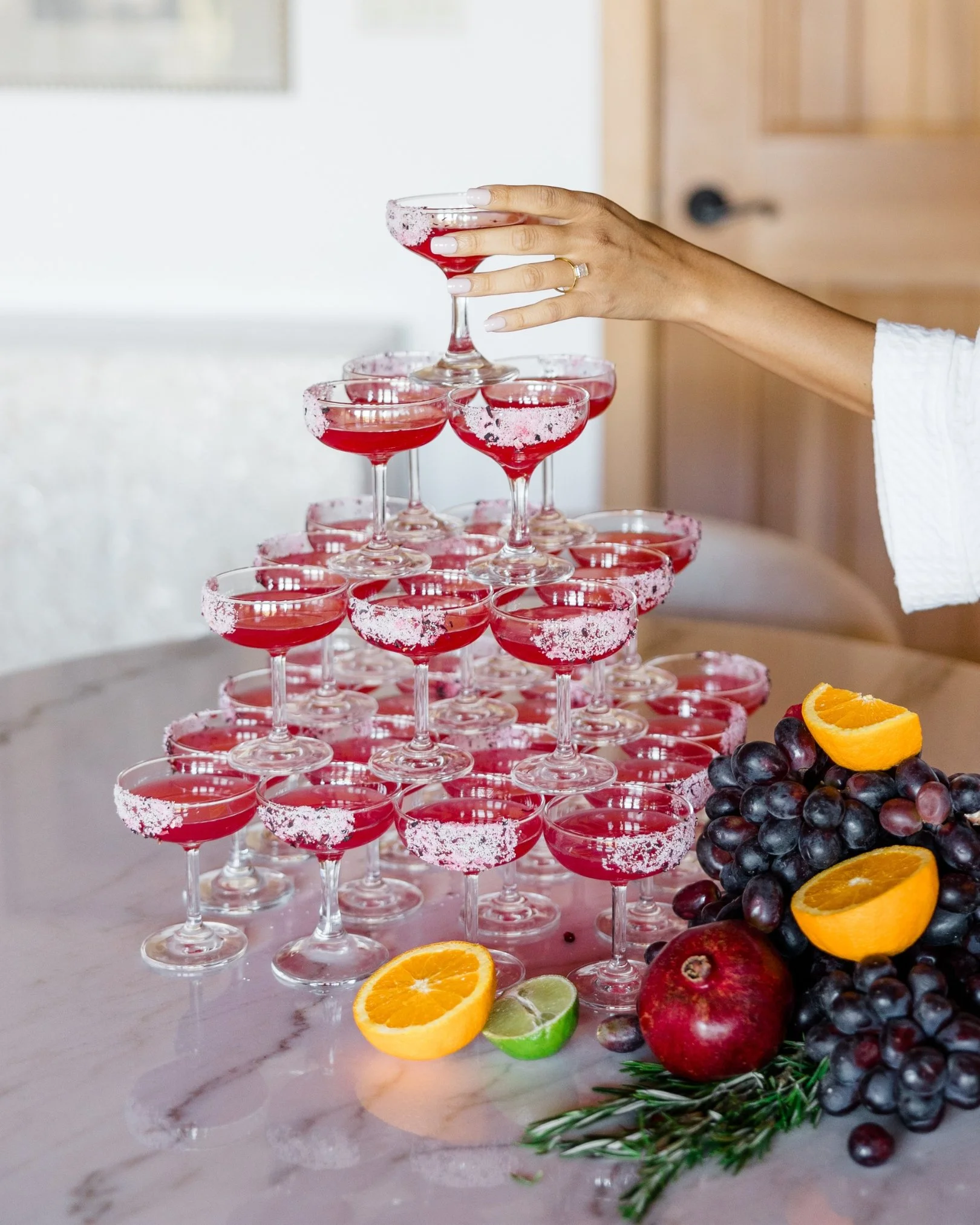 A person's hand adjusting a tiered tray of cocktails in coupe glasses with snowy rims, with fresh fruit including oranges, limes, grapes, and pomegranates on a marble surface.