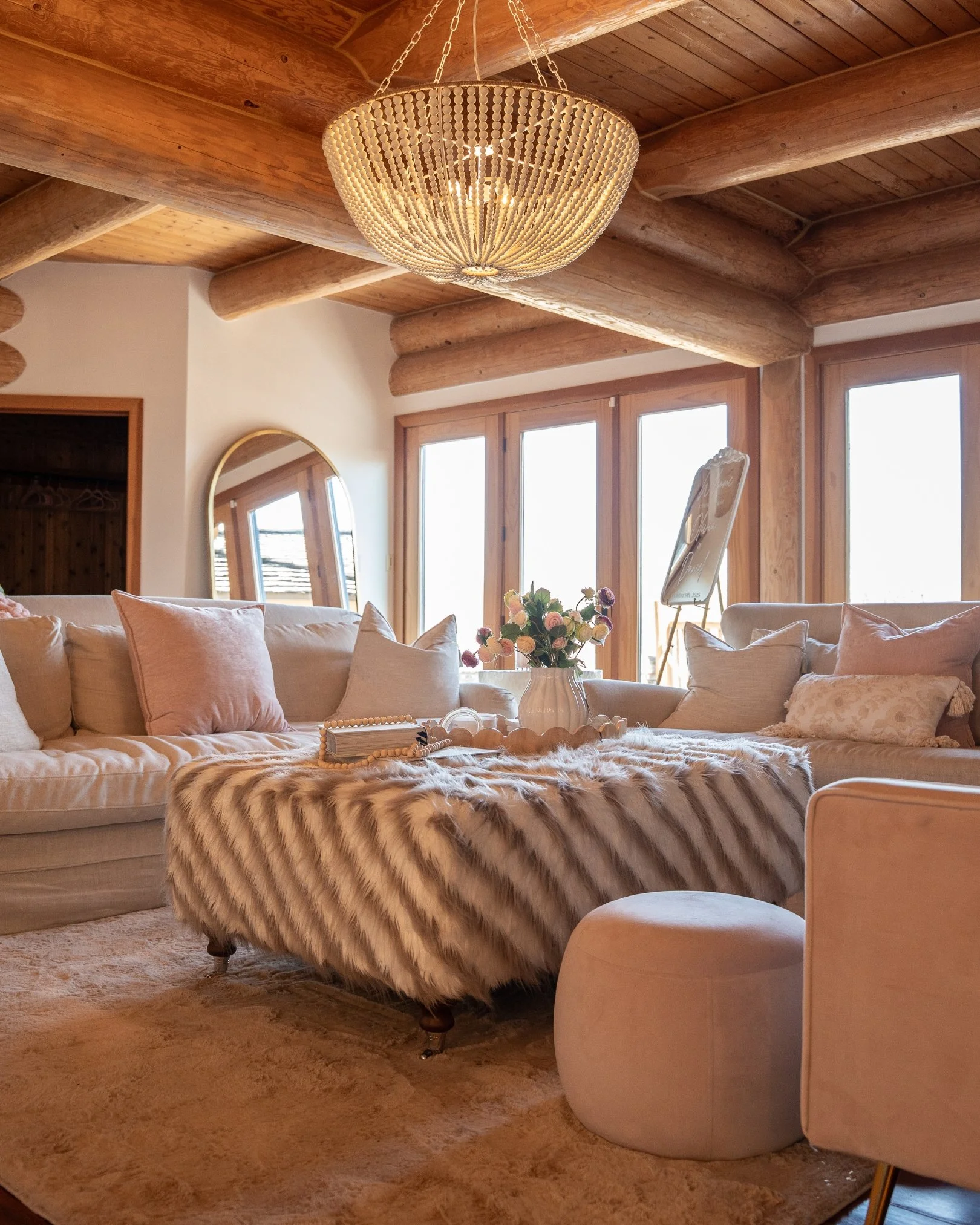 Living room with beige sofa, fur-lined ottoman, white vase with pink and purple flowers, and large windows with wooden frames. There is a wooden log ceiling with a large circular beaded chandelier and a mirror leaning against the wall