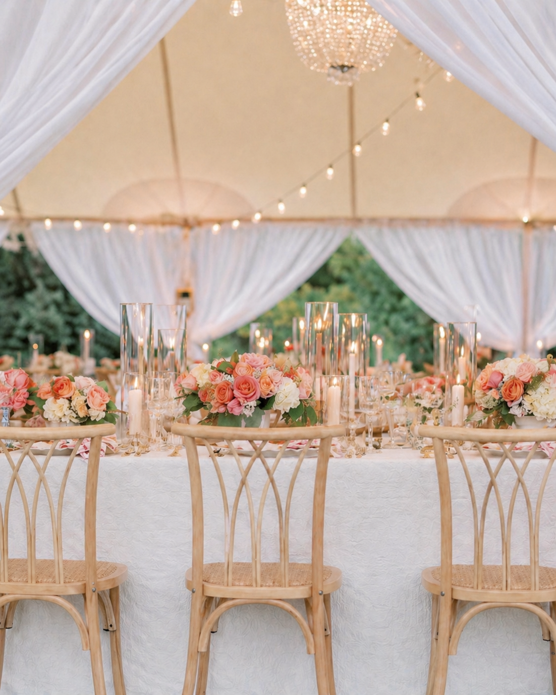 Elegant wedding reception table with floral centerpieces, candles, and clear glass vases under a decorated tent with string lights and chandeliers, surrounded by wooden chairs.