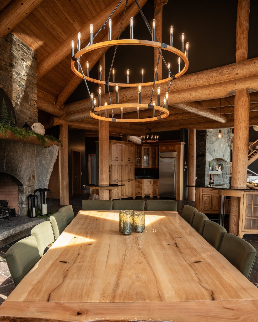 Rustic dining room featuring a large wooden table, green chairs, a double-tiered circular chandelier, exposed log beams, stone fireplace, and wooden cabinetry in the background.