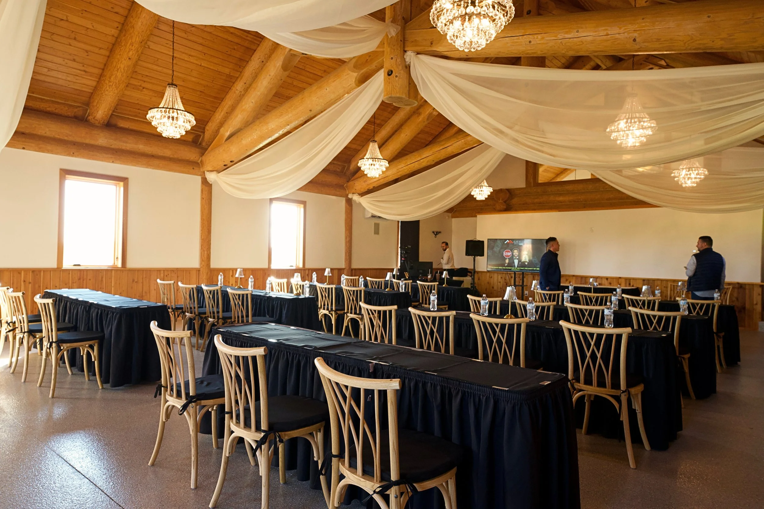 A spacious event hall with wooden walls and ceiling, decorated with sheer white fabric draped from the ceiling, chandeliers, and large windows. Tables are covered with black tablecloths and surrounded by wooden chairs with black seats. People are standing and conversing near a television screen displaying a presentation, with some water bottles placed on the tables.