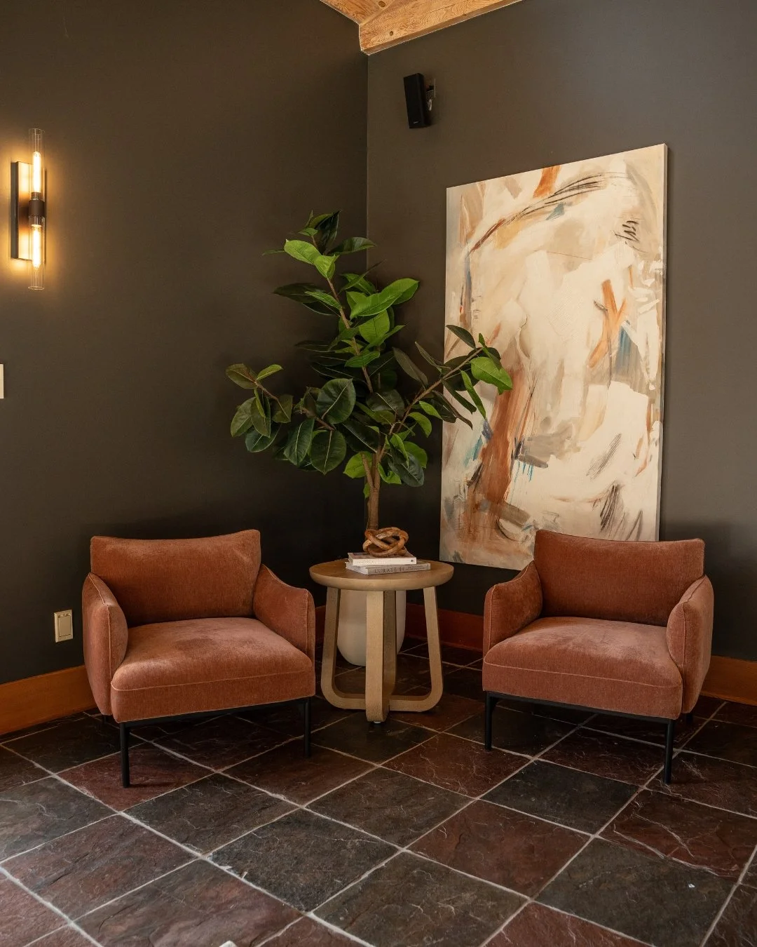 A cozy corner with two pink armchairs, a small round table with books, a large green potted plant, a modern wall light, abstract art on a dark gray wall, and a wooden ceiling.