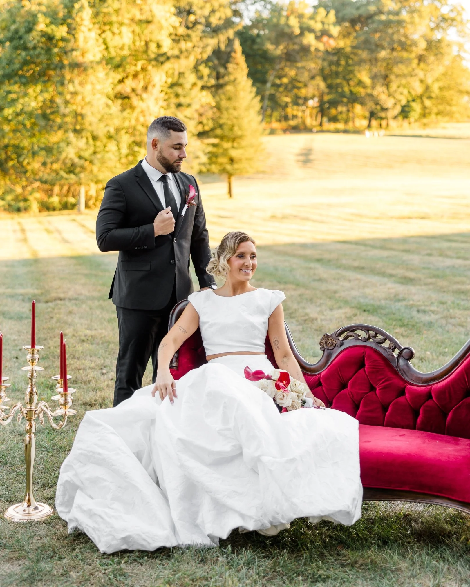 A woman in a white dress sitting on an ornate red velvet sofa outdoors, holding a bouquet of roses, with a man in a black suit and tie standing beside her, touching her shoulder, on a grassy lawn with trees and a lake in the background during sunset.