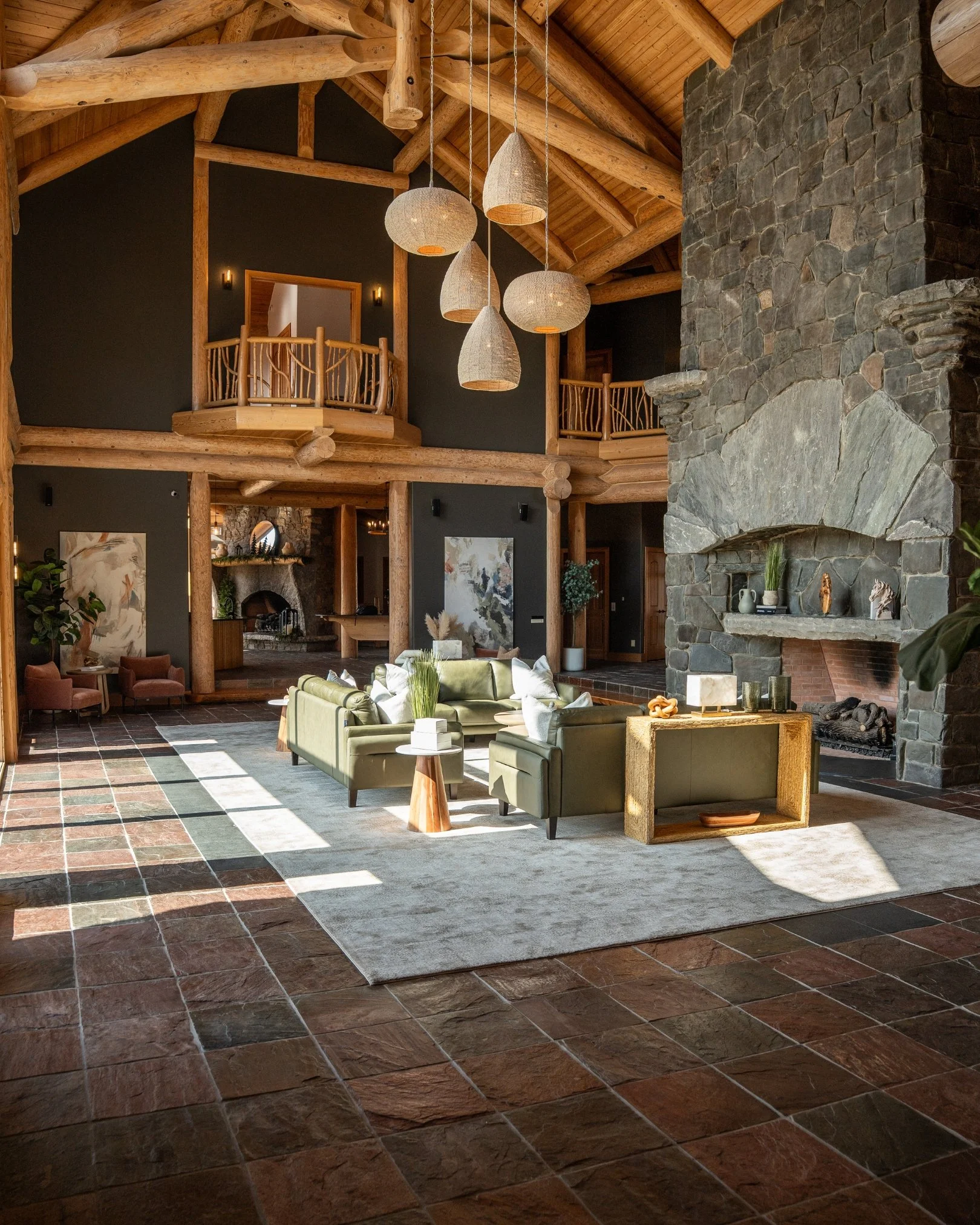Interior of a rustic living room featuring a large stone fireplace, wooden beams and trim, modern green sofas arranged around a coffee table, and hanging pendant lights in woodmont manor