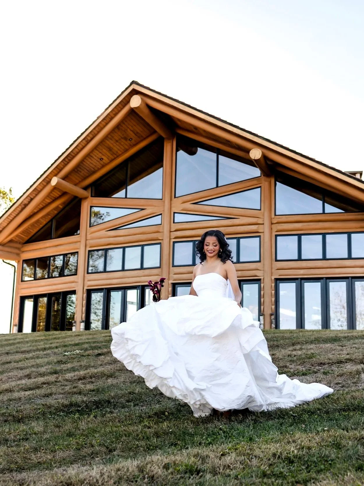 A bride in a white wedding gown sitting on the grass in front of a large wooden house with glass windows.