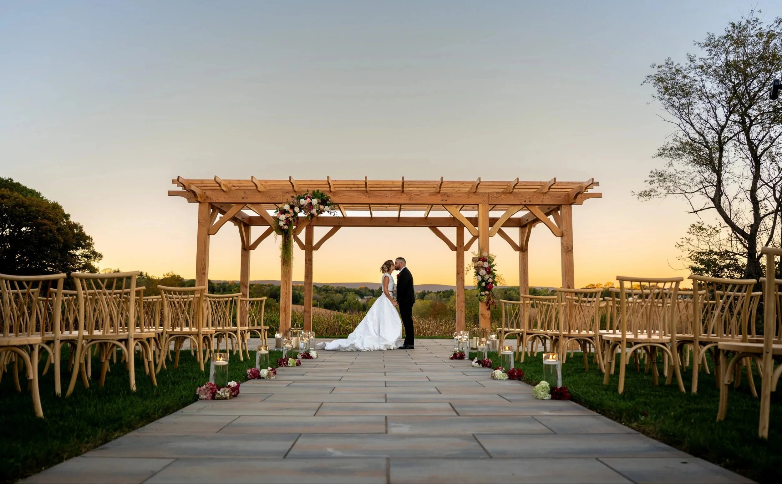 A bride and groom standing under a wooden wedding arch decorated with flowers, at sunset outdoors.