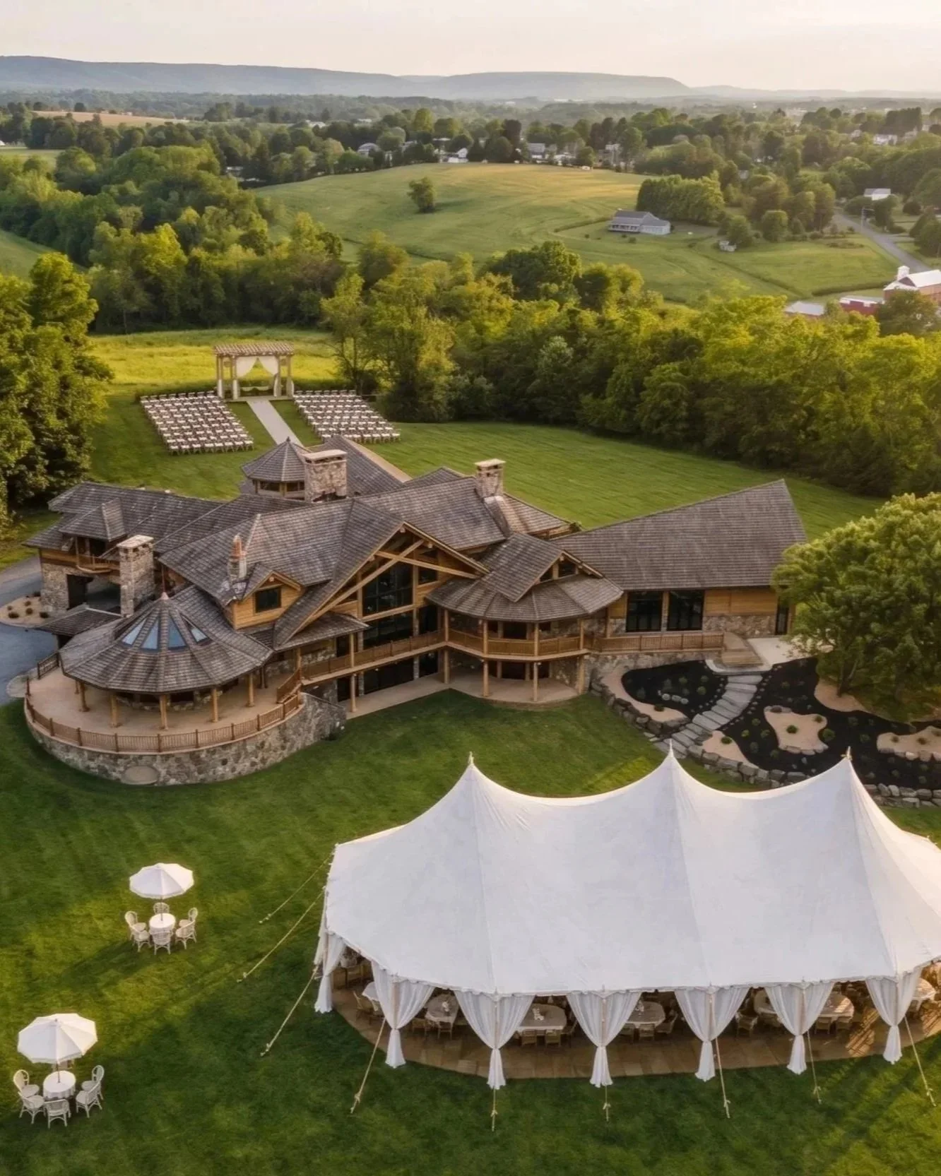 A large estate with a modern house, outdoor tent, and picnic tables in a lush green landscape with trees and rolling hills in distance.