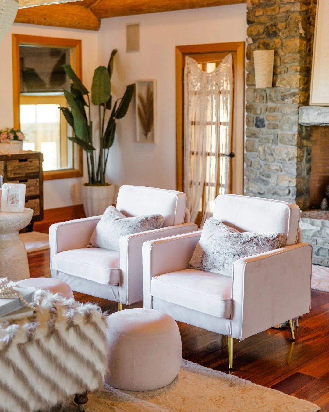 A cozy living room with two white armchairs with pillows, a beige ottoman, a stone fireplace, a large potted plant, a window, a glass door with sheer curtains, and wooden ceiling beams.