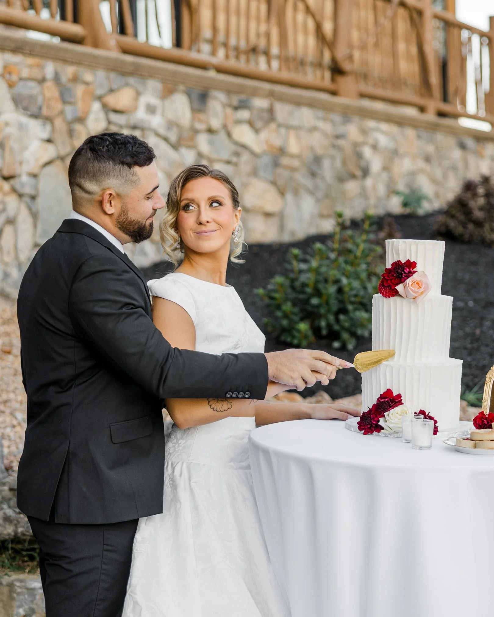A bride and groom at a wedding cake cutting ceremony outdoors. The bride is holding a knife, smiling at the groom, who is wearing a black suit, against a stone wall background with a wooden railing above.