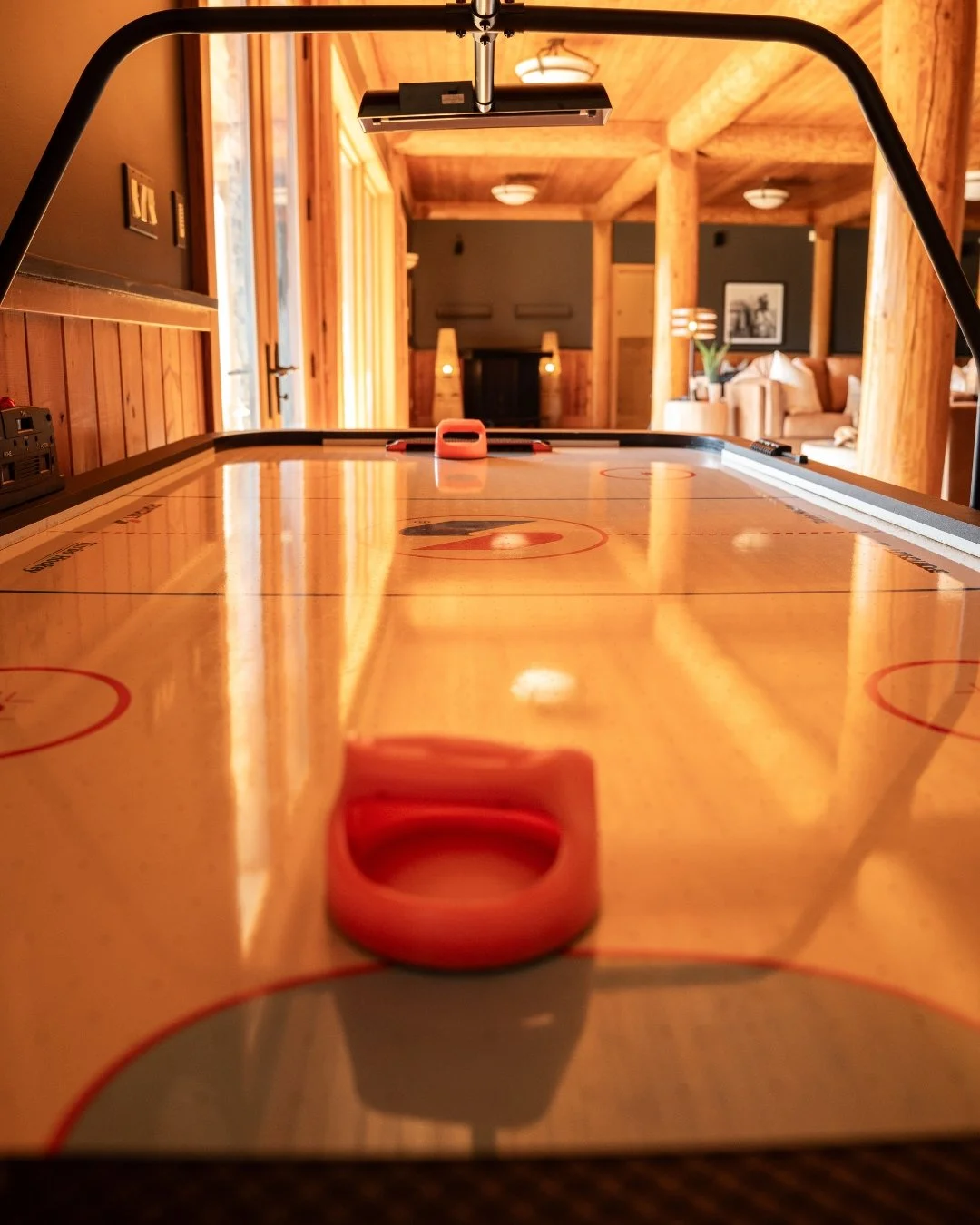 An air hockey table with a shiny wooden surface in a cozy, wood-paneled room, with a background of living room furniture, lamps, and wood beams.