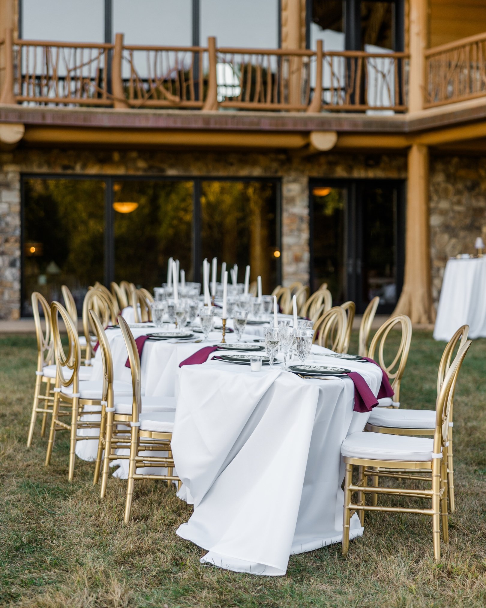 Outdoor wedding reception setup with a long table covered in a white tablecloth, decorated with glassware and purple napkins, surrounded by gold chairs on a grassy area, with a rustic building with large windows and a wooden balcony in the background.