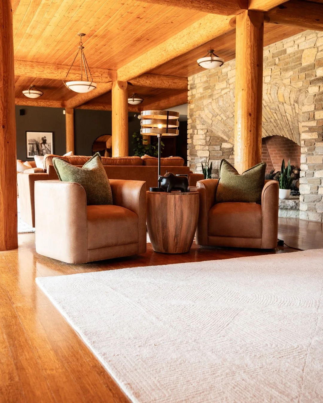 Cozy living room with wooden ceiling, brick fireplace, leather armchairs, and a table with a lamp and decorative animal figure.