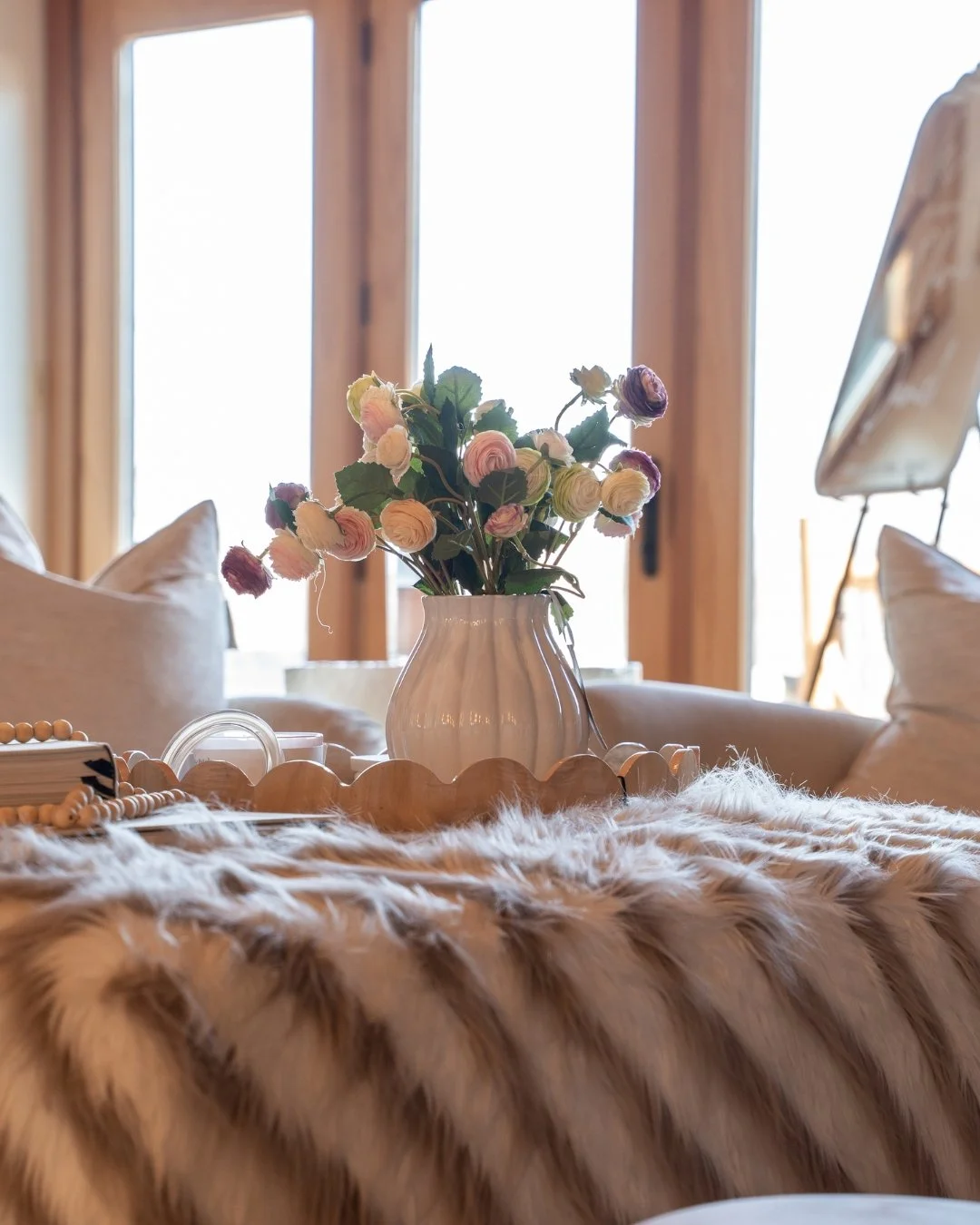 A white vase filled with pastel-colored ranunculus flowers, placed on a table with a faux fur throw. In the background, there are large windows with wooden frames and beige cushions on a sofa.