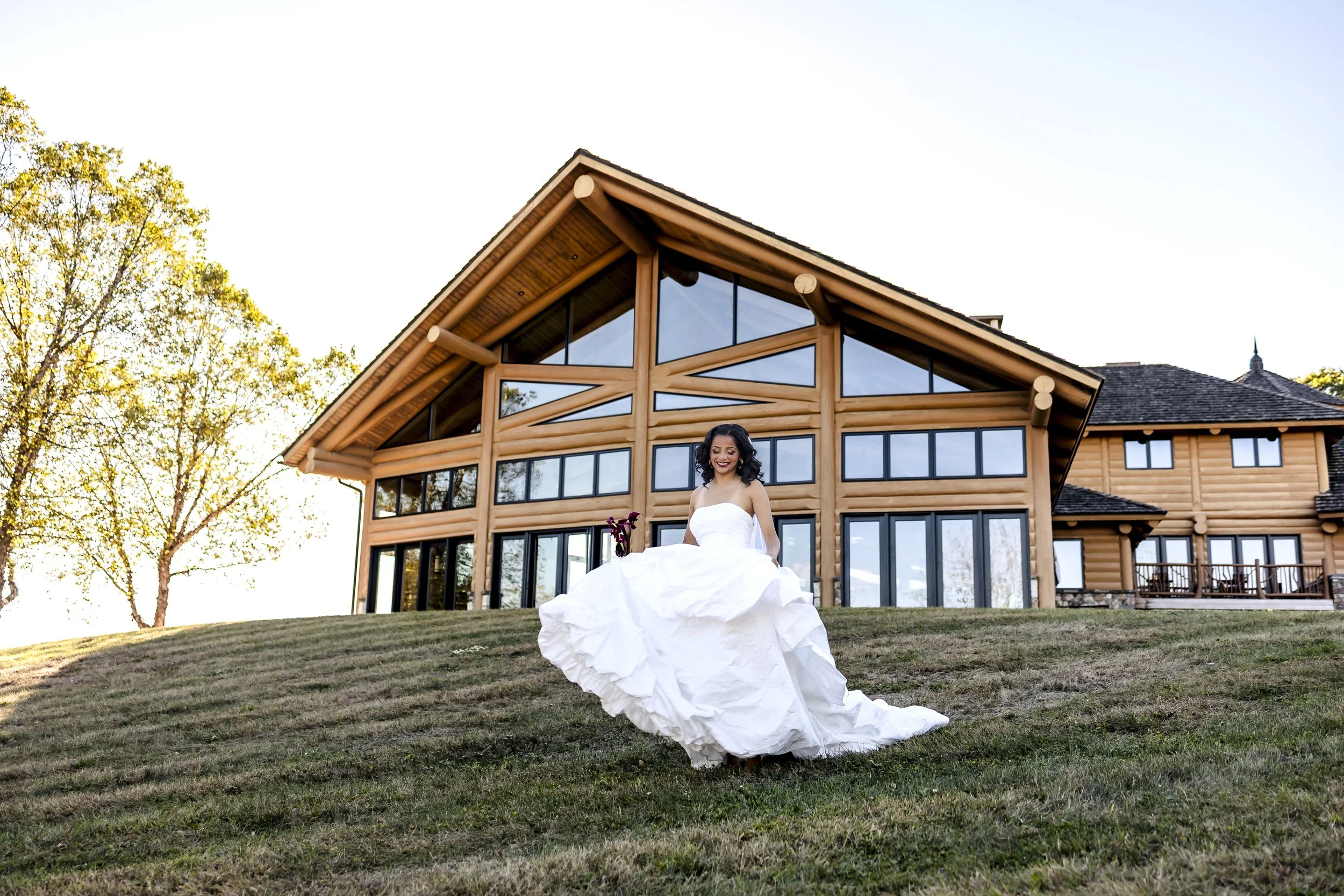 A woman in a white wedding dress sitting on a grassy lawn in front of a large wooden house with many windows and a sloped roof, surrounded by trees with green leaves.