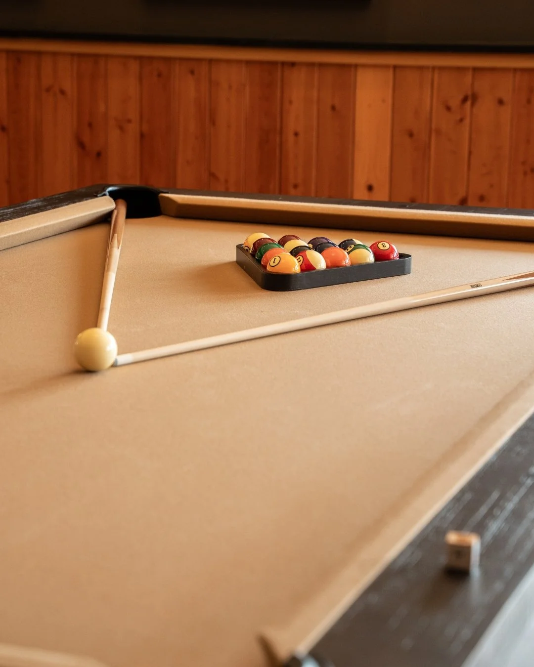 Pool table with a cue stick resting on the edge, a rack of billiard balls in the corner, and a wooden paneled wall in the background.