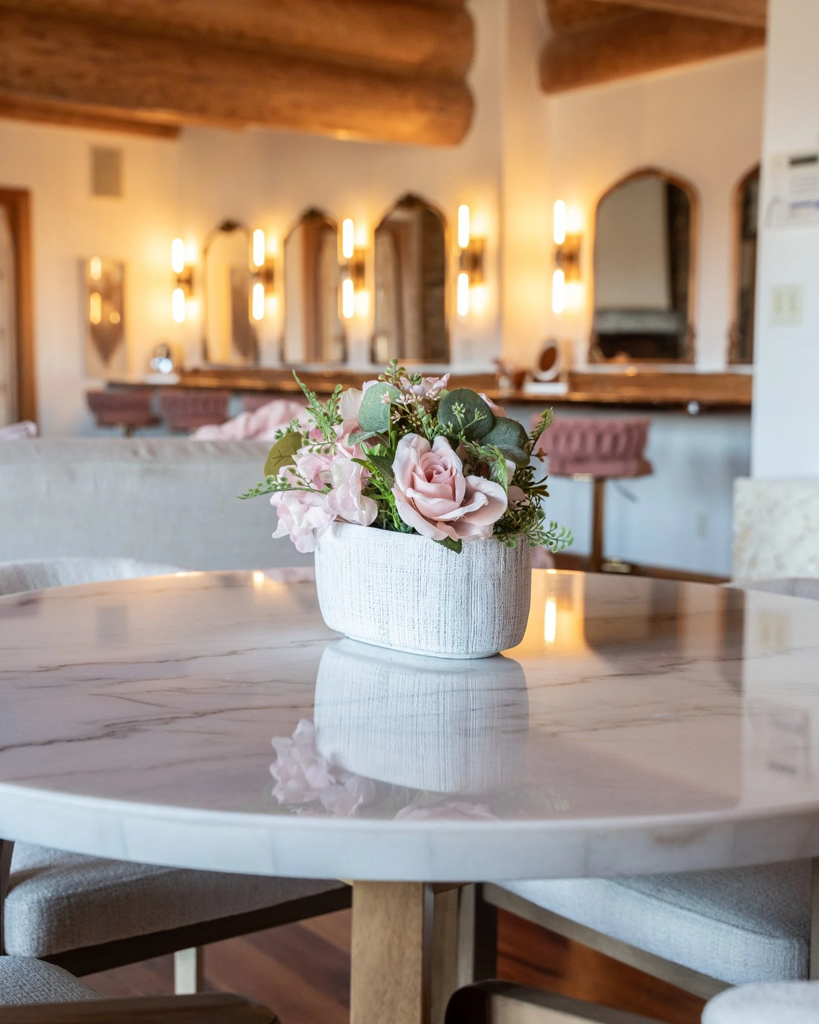 A white textured flowerpot with pink roses, eucalyptus, and other greenery on a marble table in a cozy room with wooden accents and soft lighting.