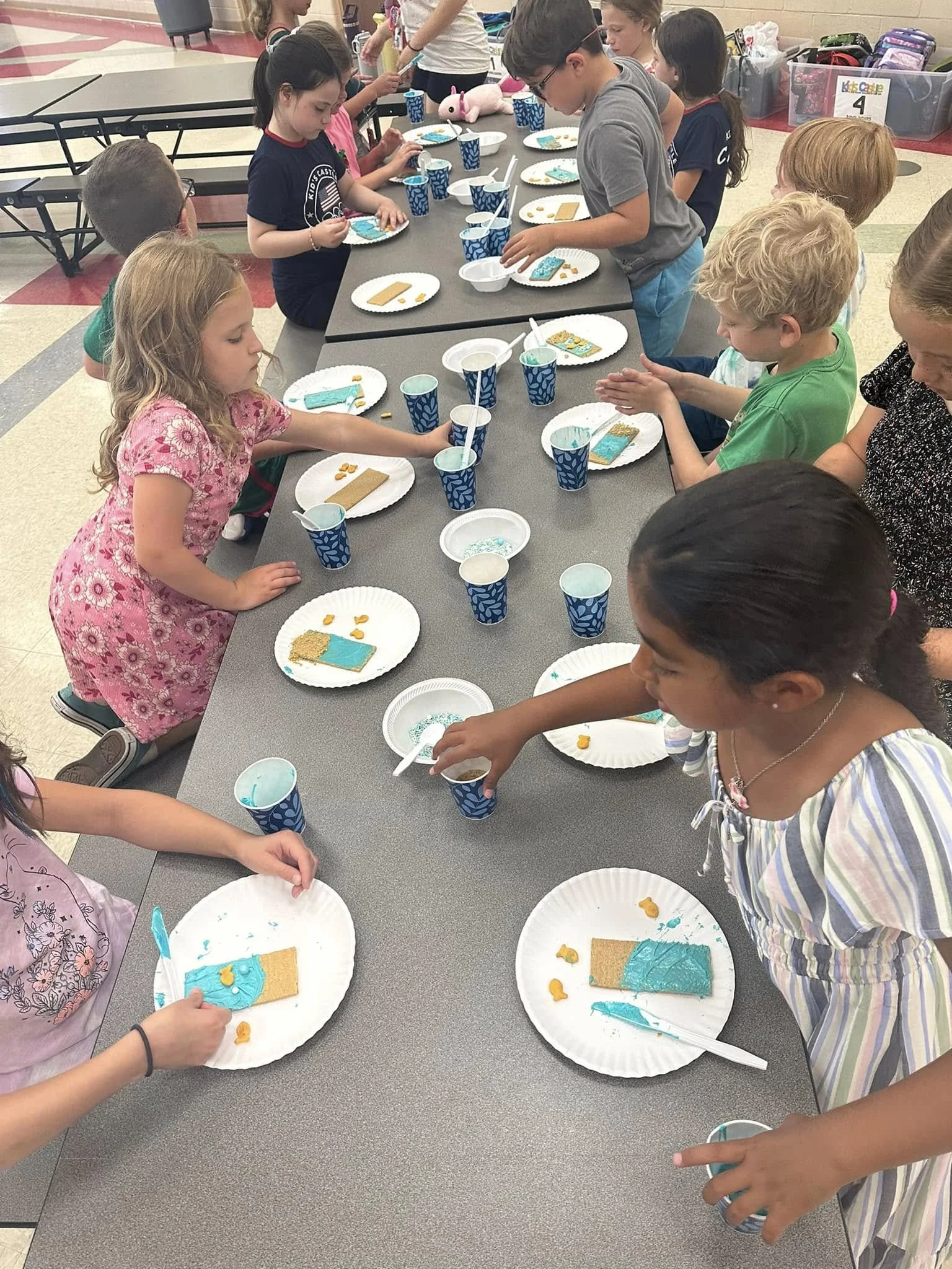 Children at a table decorating cookies with blue icing and yellow candies in a classroom or cafeteria setting.