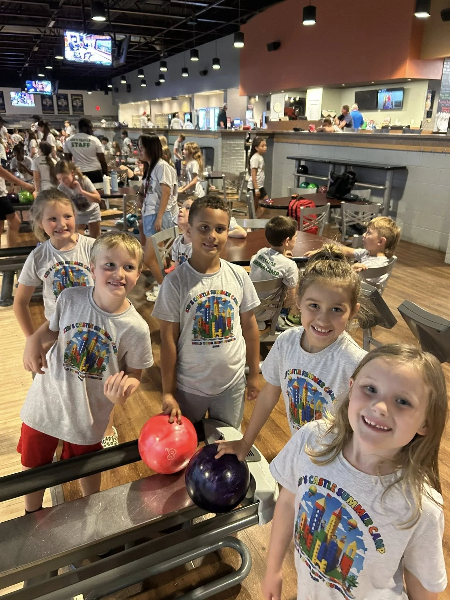 Children at a bowling alley, some holding colorful bowling balls, wearing T-shirts with a cityscape design and the words 'Castle Summer Camp,' with other kids and adults in the background.