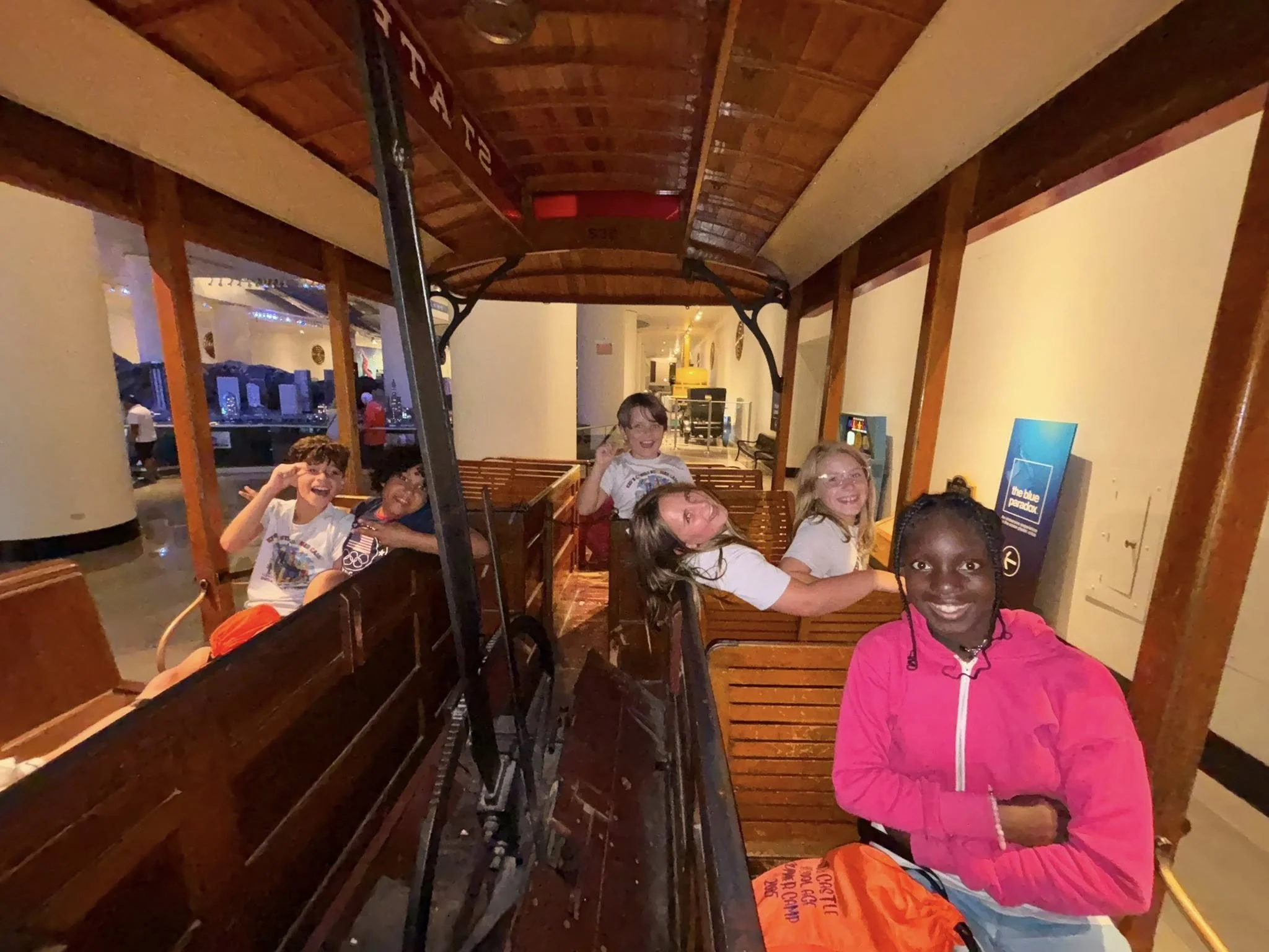 Group of smiling children sitting inside a vintage train carriage at an indoor museum or exhibit, with wooden benches and a wooden ceiling.