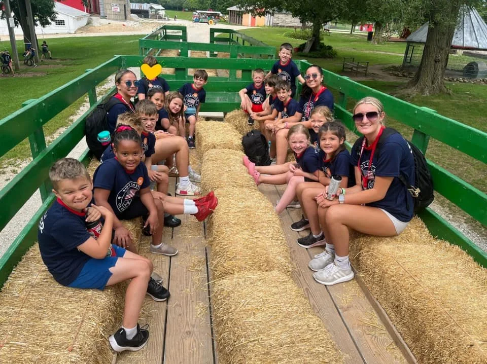 Group of children and two adults sitting on hay bales inside a wooden wagon at an outdoor park.