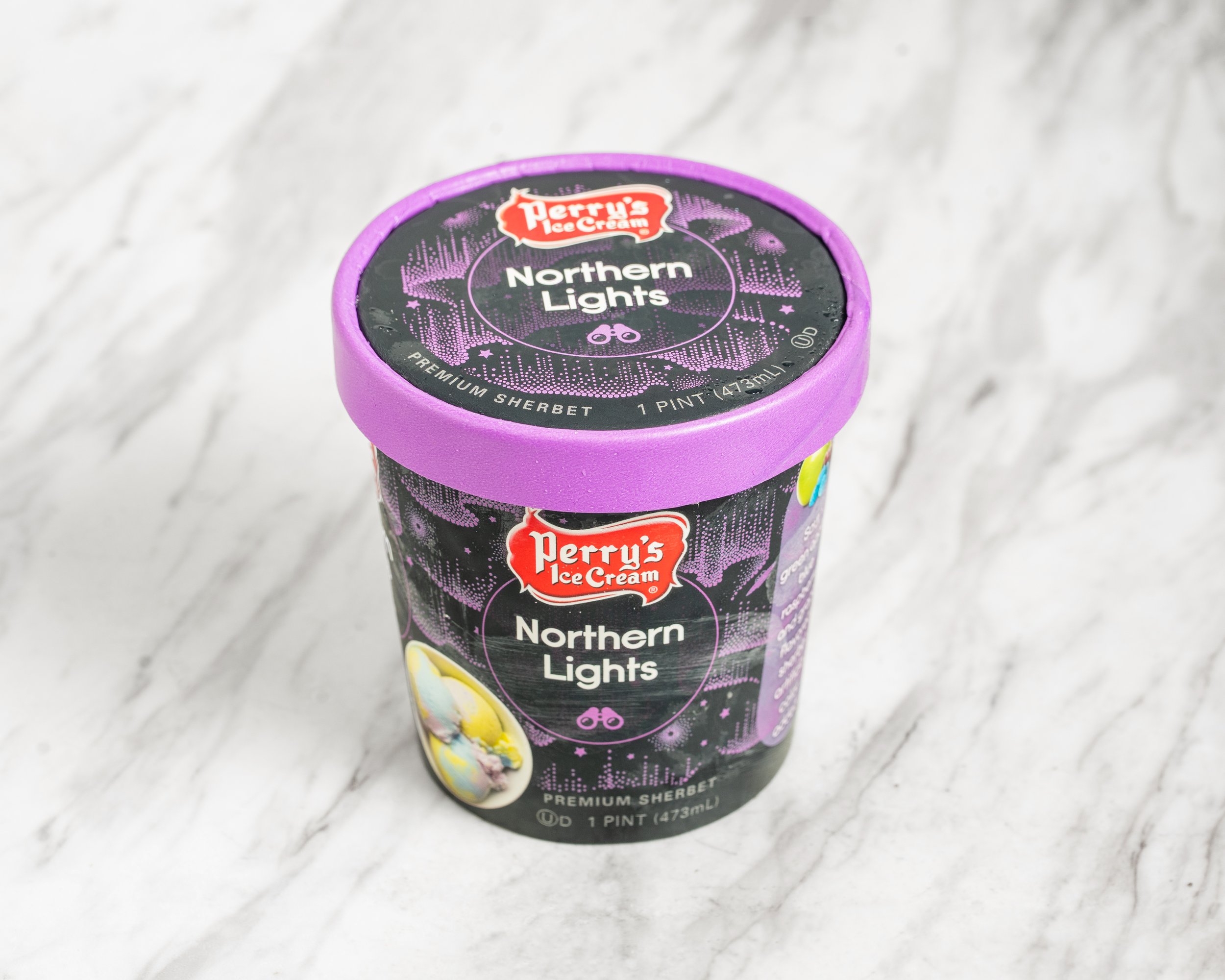 A pint of Perry's Ice Cream Northern Lights flavor with a purple lid on a marble surface.