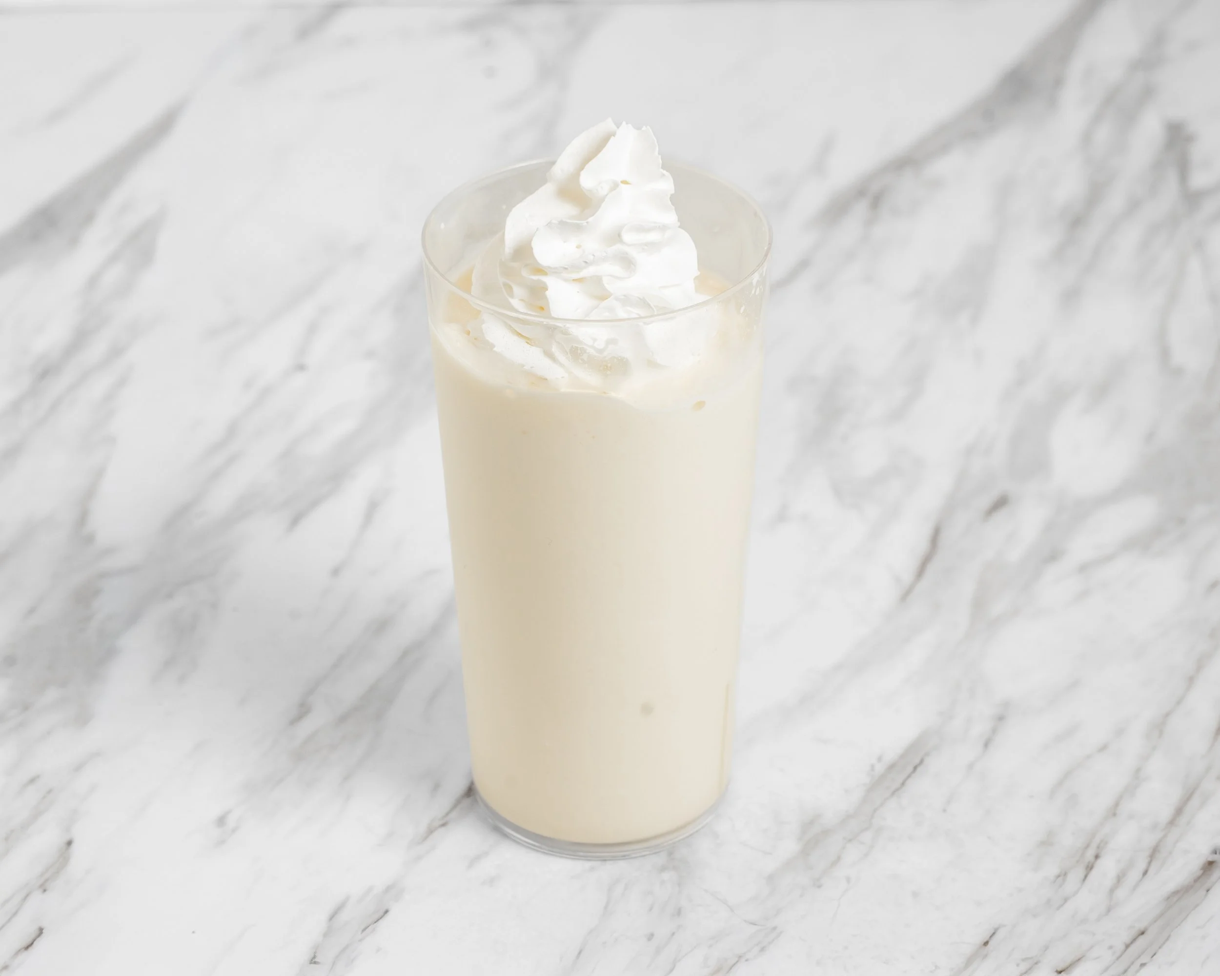 A tall glass of vanilla milkshake topped with whipped cream on a white marble surface.