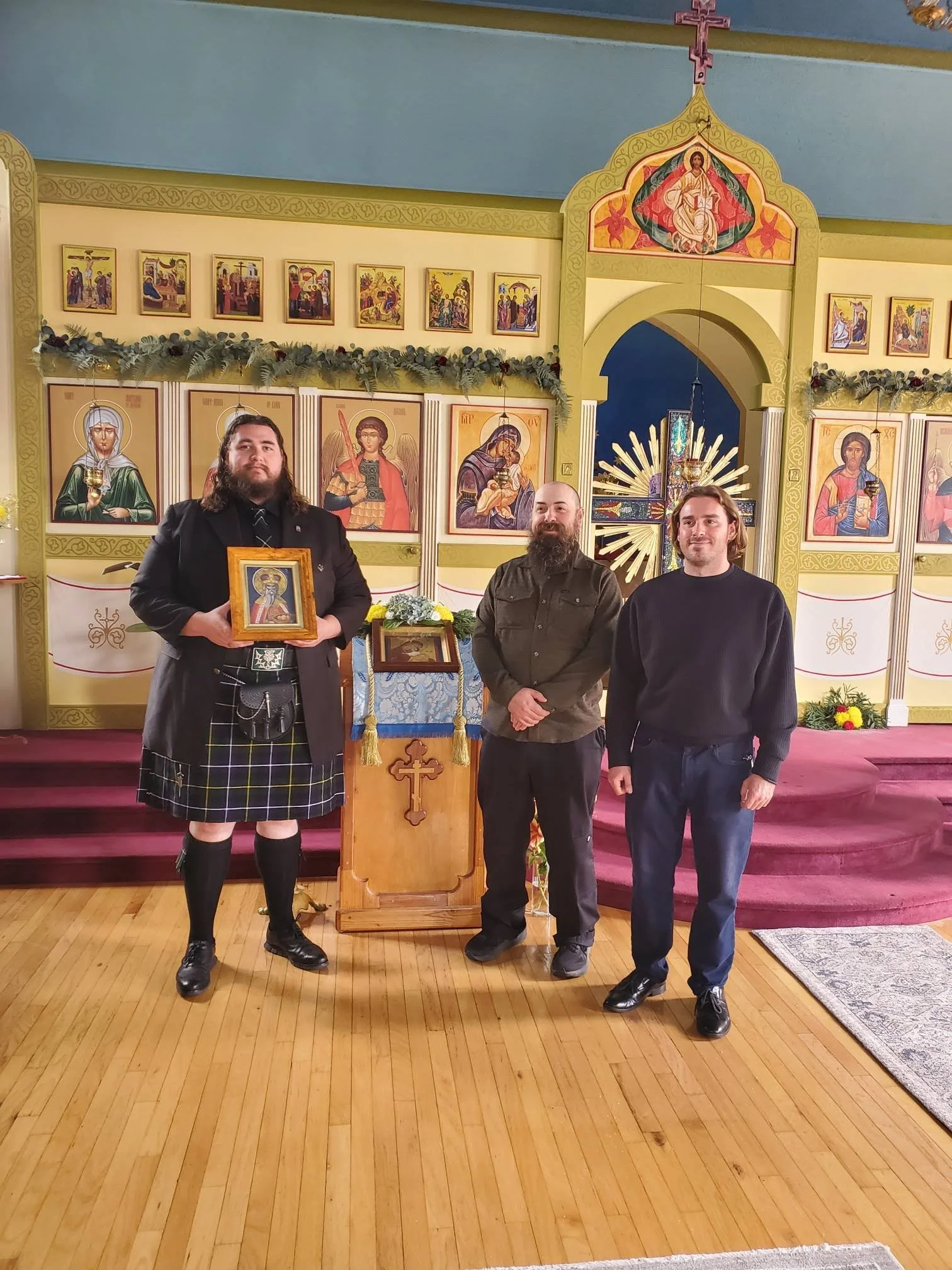 Three men stand inside an ornately decorated church or chapel with religious icons and artwork on the walls. One man holds a framed religious icon, and the others stand beside him. The interior features gold accents, floral decorations, and a wooden podium with a cross in front of the icons.