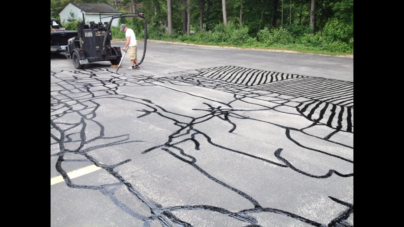 Person working on a large black chalk drawing on an empty parking lot with trees in the background.
