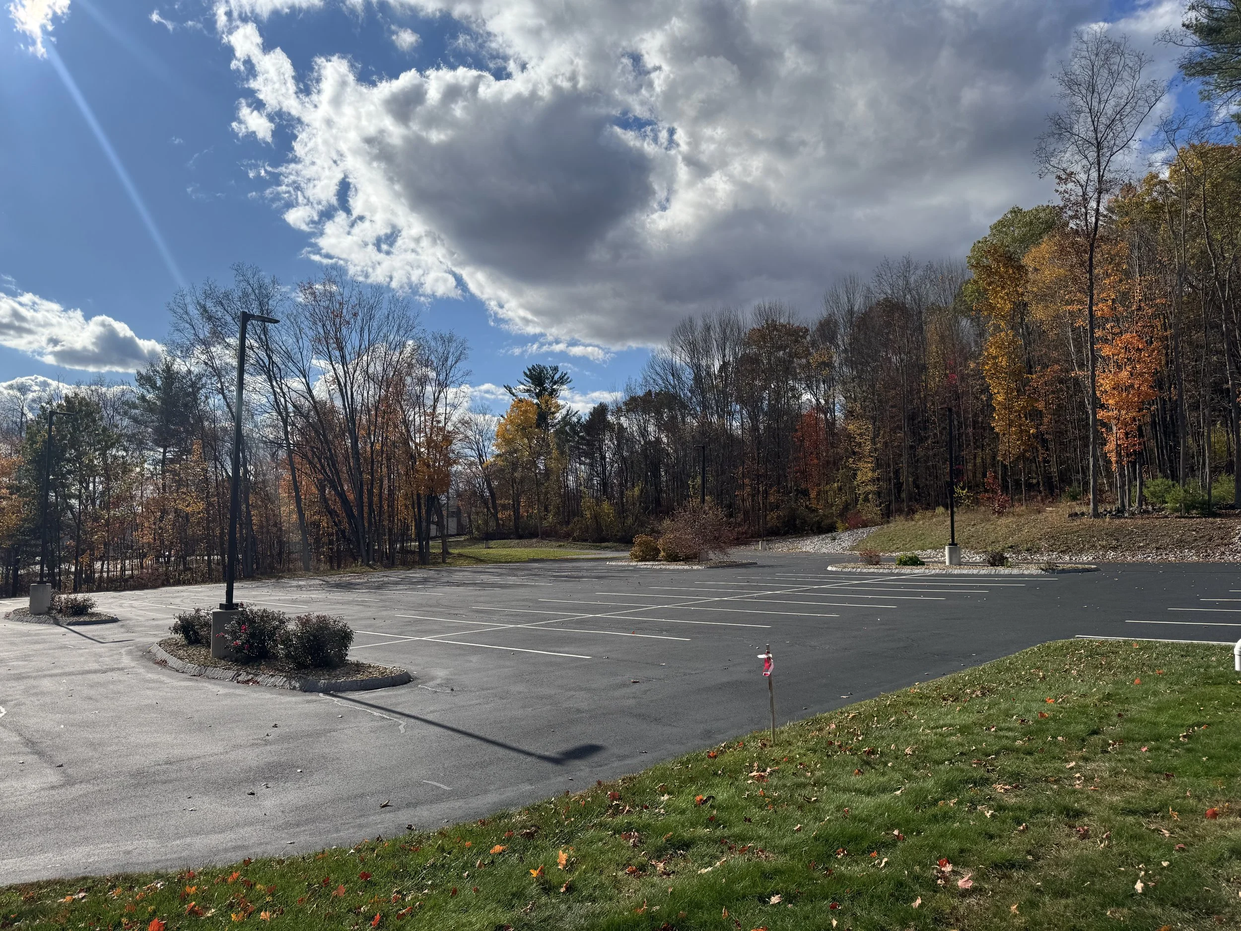 Empty parking lot surrounded by trees with autumn leaves, under a partly cloudy sky with the sun shining.