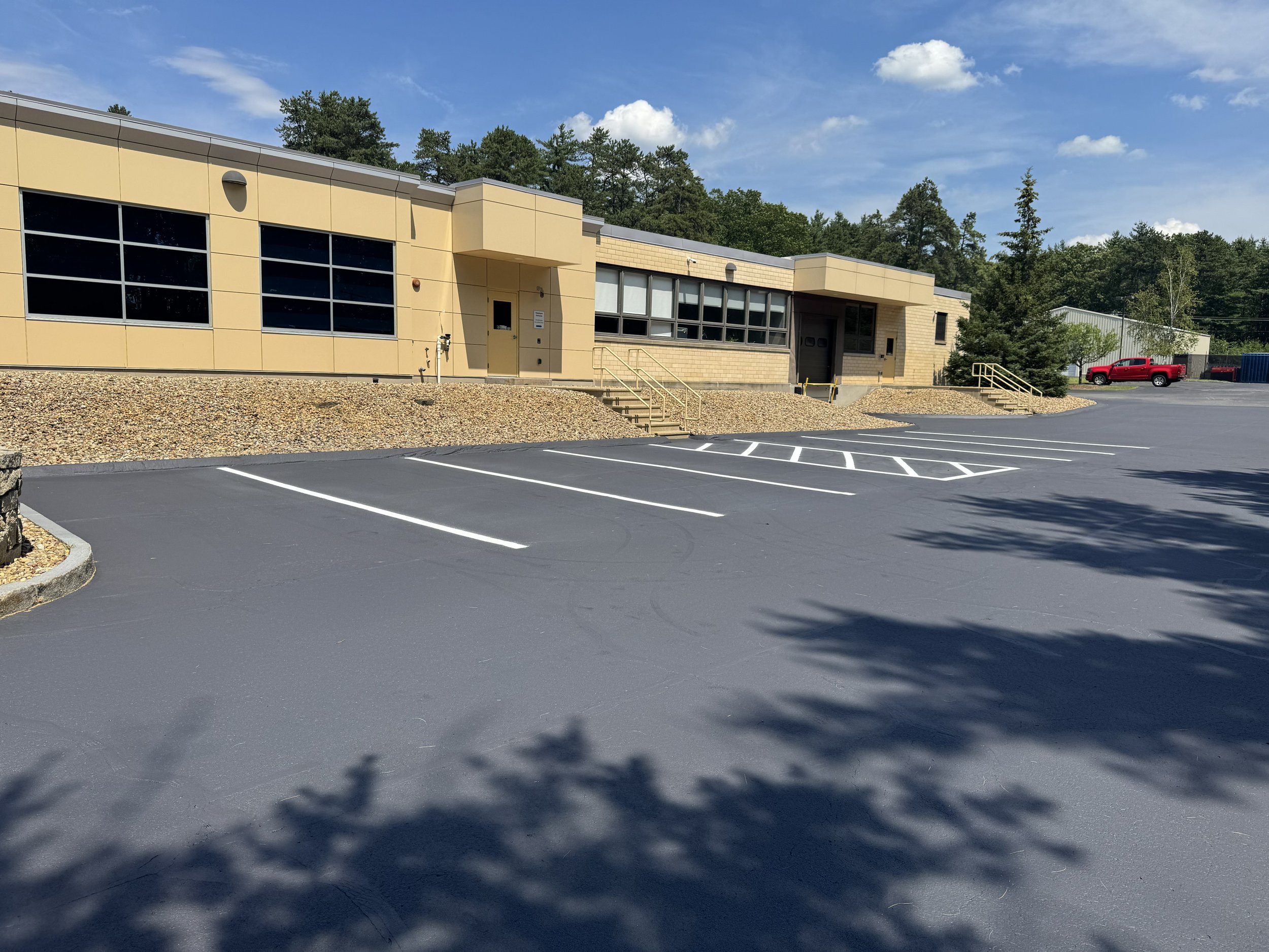 Empty parking lot in front of a beige commercial building with large windows, stairs leading to entrance, and a red pickup truck parked to the right.