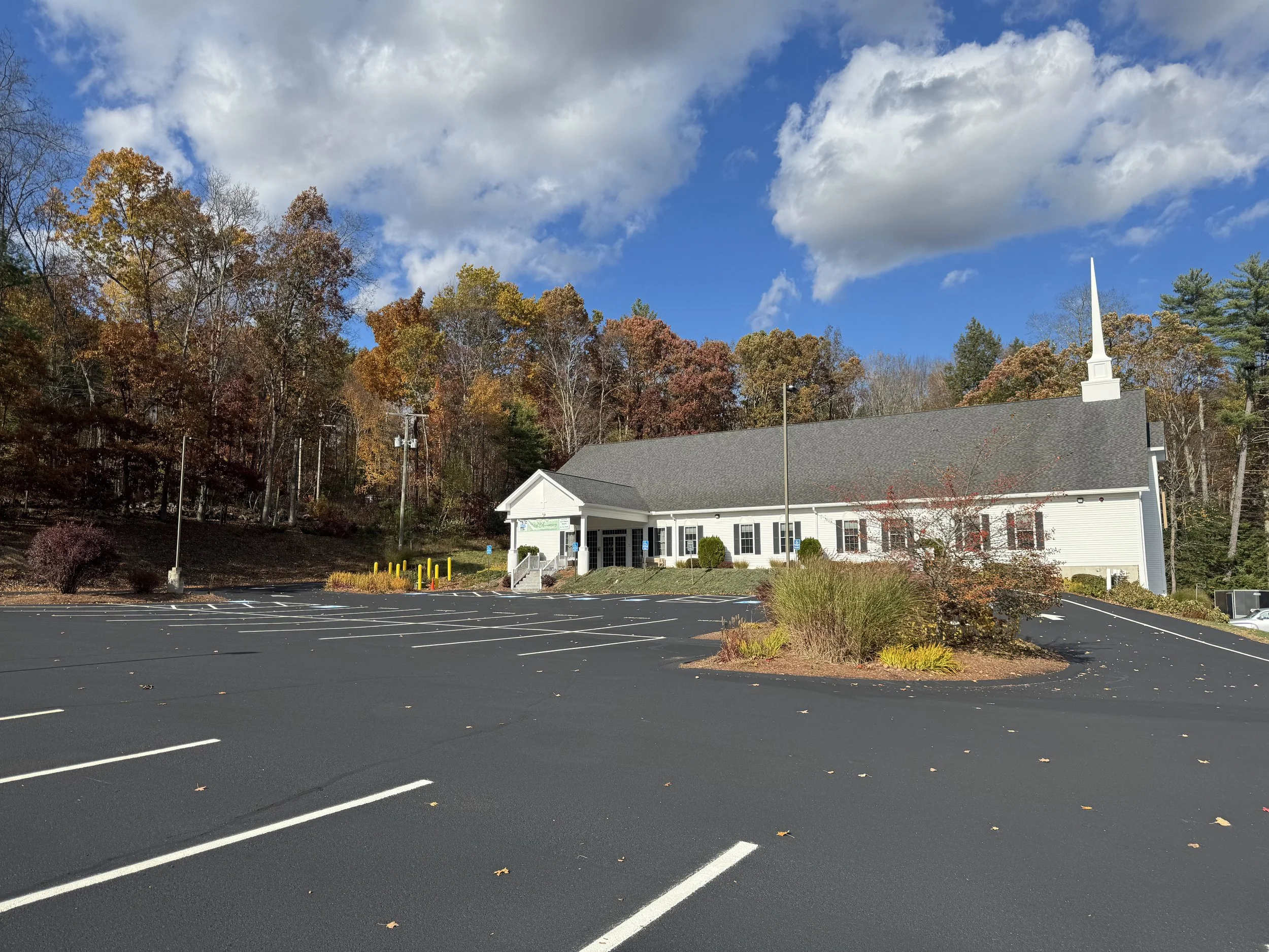 A church with a white exterior and tall steeple, surrounded by trees with fall foliage, and an empty parking lot in the foreground.