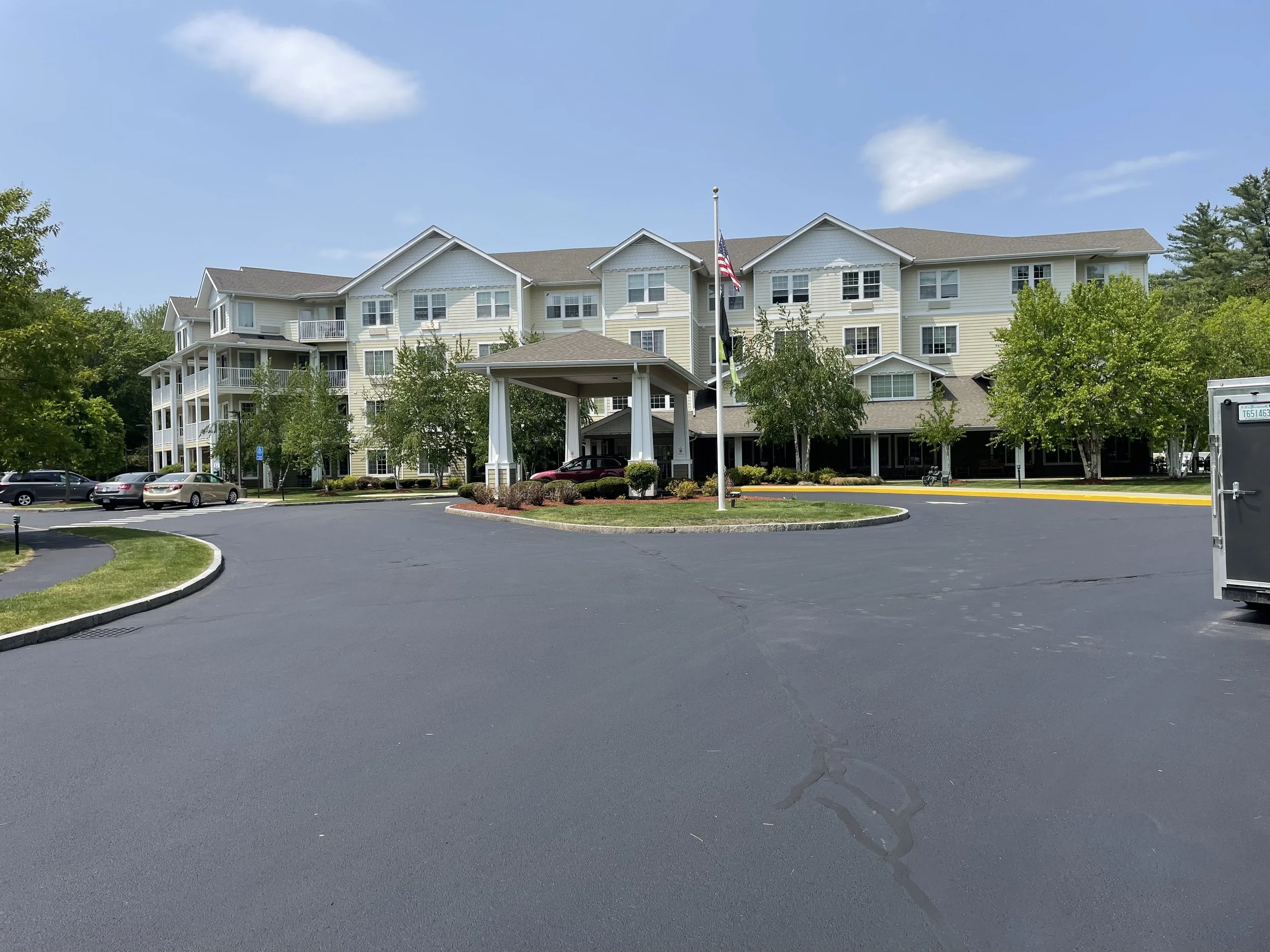 A multi-story residential building with a covered entrance and flags in front, surrounded by trees, with a driveway and parked cars in the foreground.