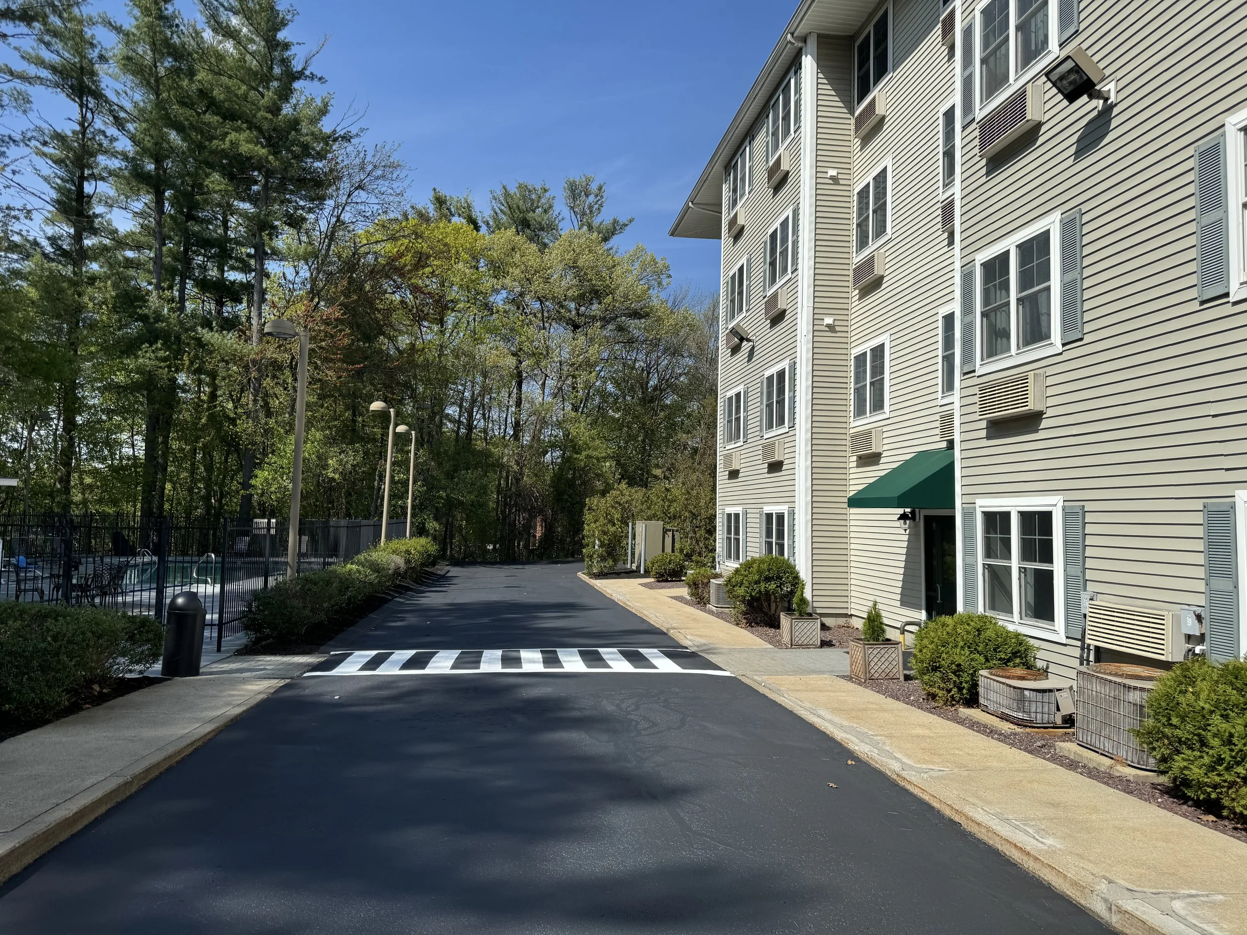 A residential apartment complex with a paved driveway, apartment building on the right, and a swimming pool with chairs on the left, surrounded by trees and a fence.