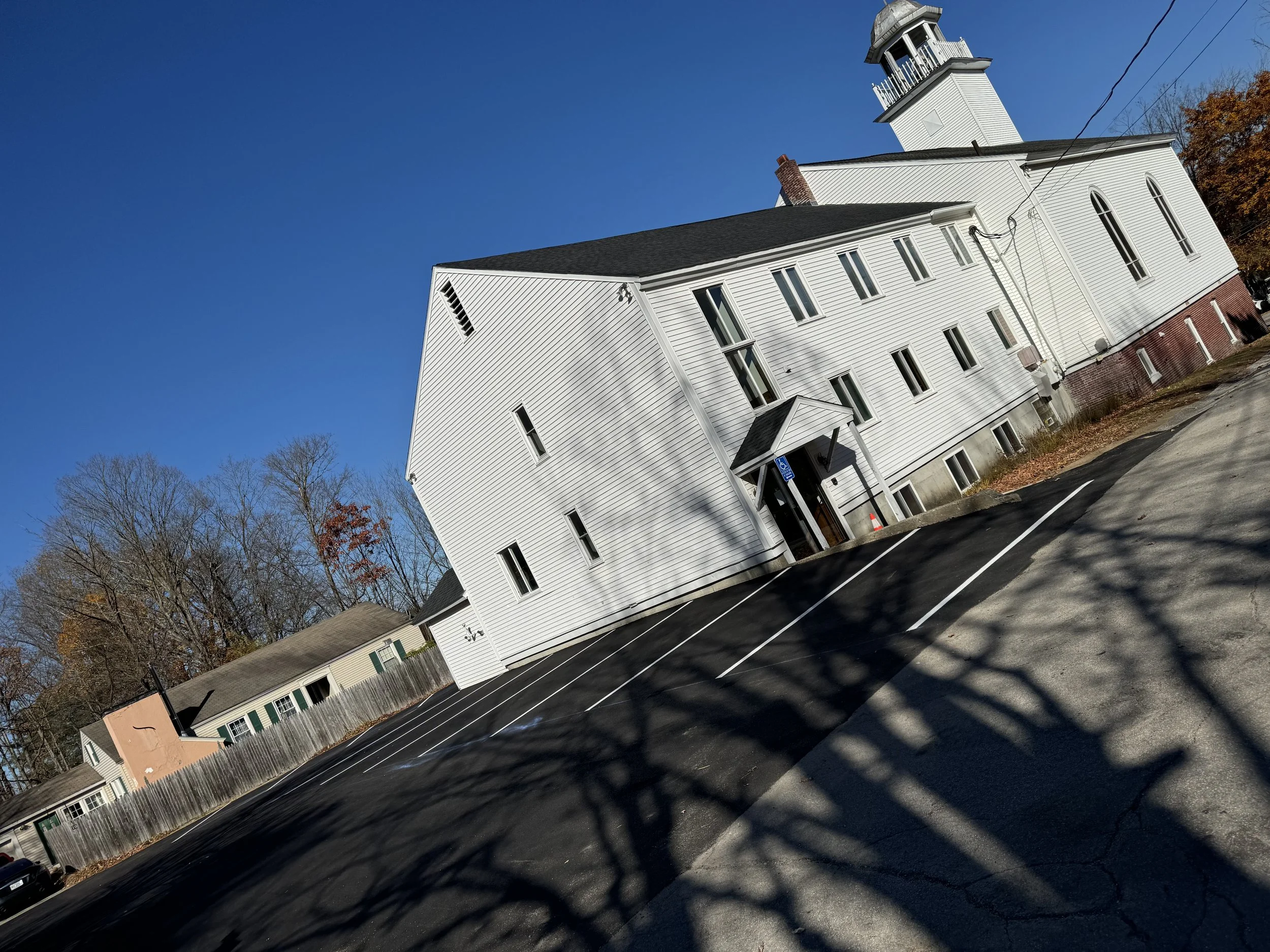 White church with steeple under a clear blue sky, surrounded by leafless trees, with an empty parking lot in the foreground.