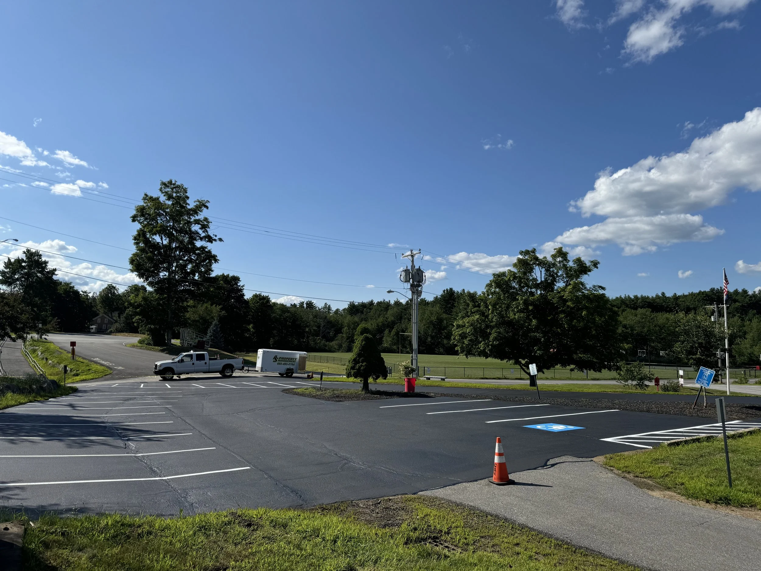 A parking lot with designated handicap parking space, traffic cone, trees, a truck and trailer, utility pole, grassy area, and a field with a basketball hoop in the background under a bright blue sky with scattered clouds.