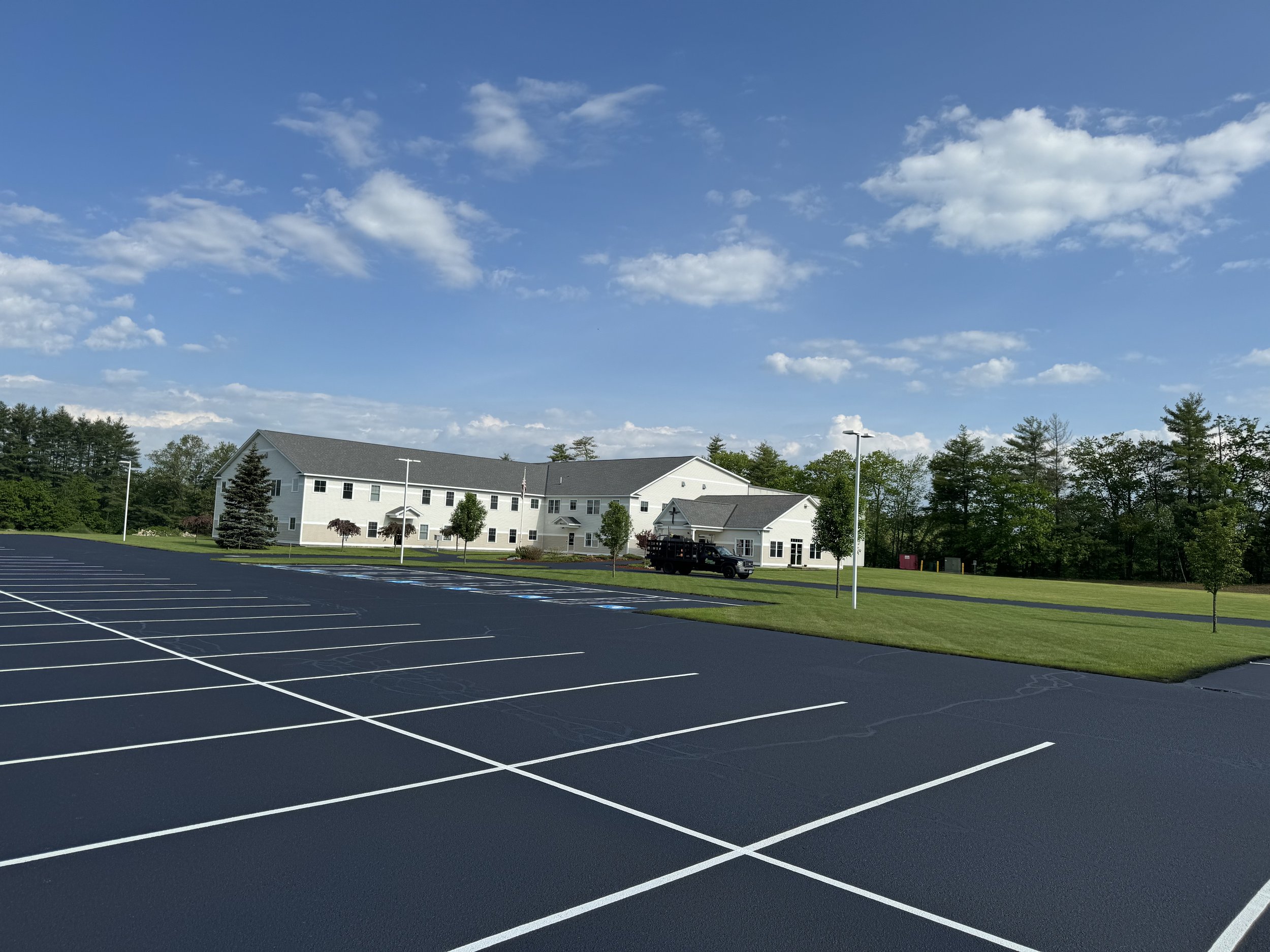 A large, mostly empty parking lot with painted white lines and a few parking spaces marked for accessible parking. In the background, there is a white multi-story building, surrounded by green grass and small trees, with a blue sky and scattered clouds overhead.