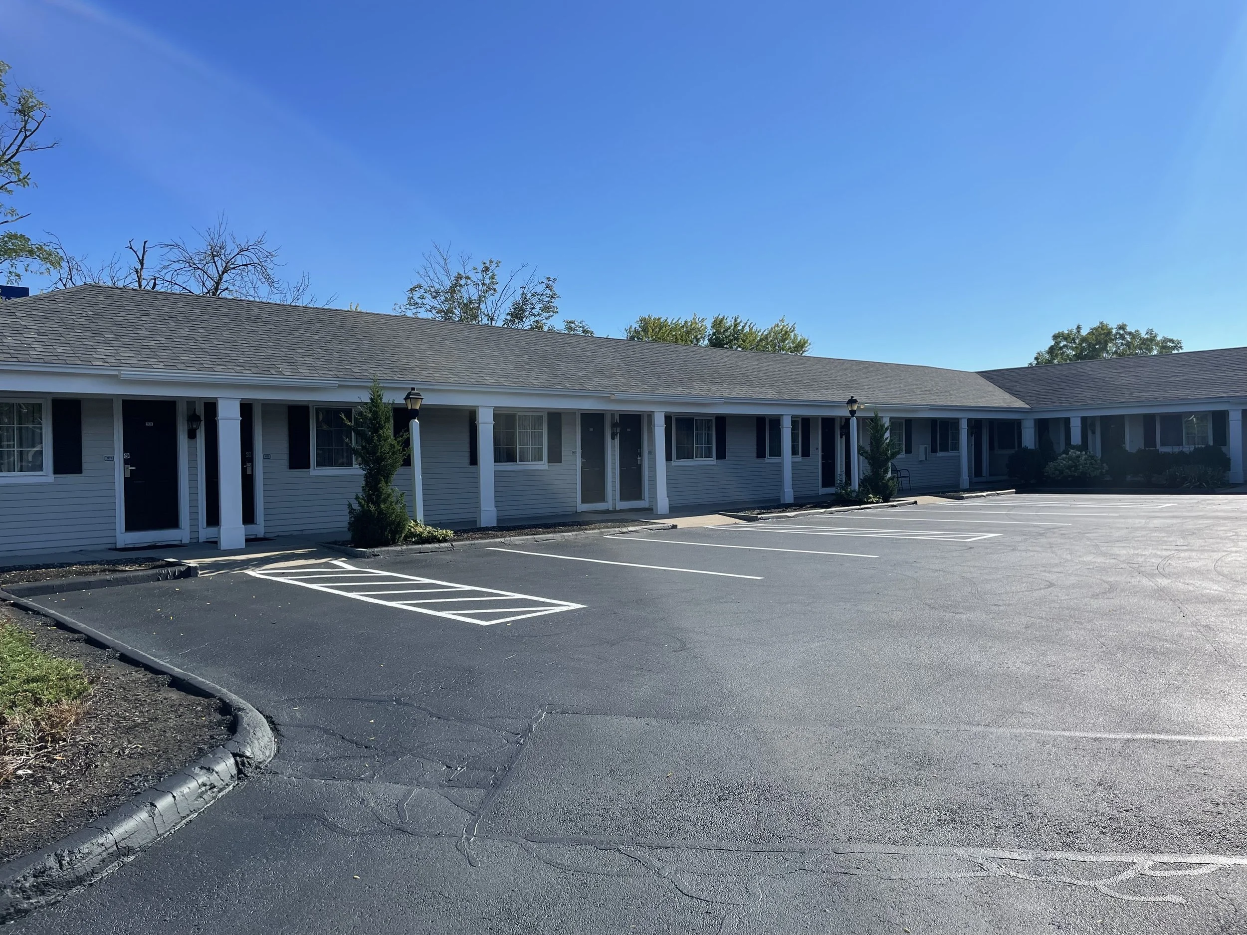 Exterior view of a single-story apartment complex with a parking lot. The building has gray siding, black shutters, and white columns, surrounded by a few small trees. The parking lot has several unoccupied spaces and is under a clear blue sky.