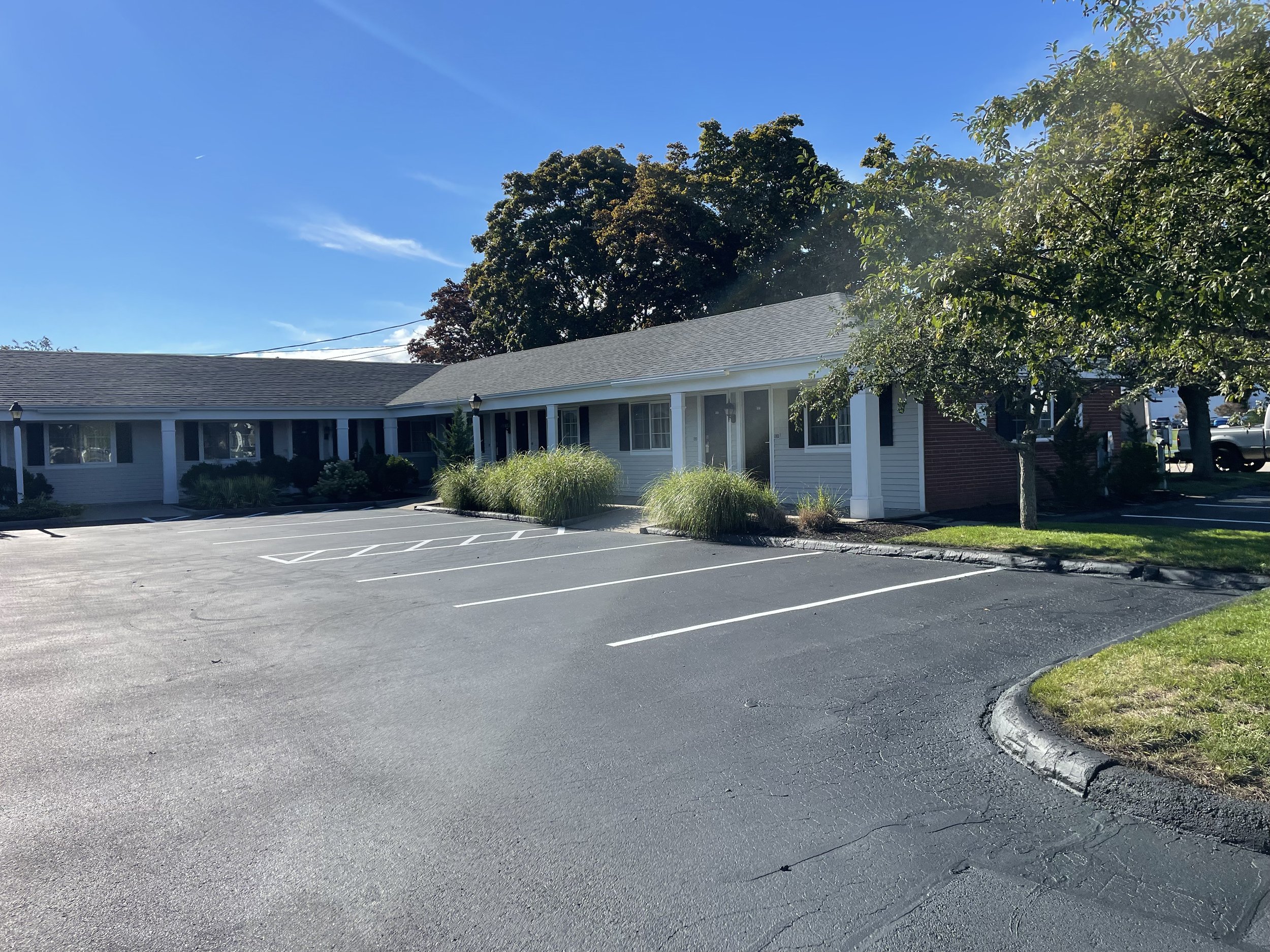 A single-story commercial building with white siding, surrounded by a parking lot with empty parking spaces. There are trees and shrubs in front of the building, and the sky is clear with sunlight shining down.