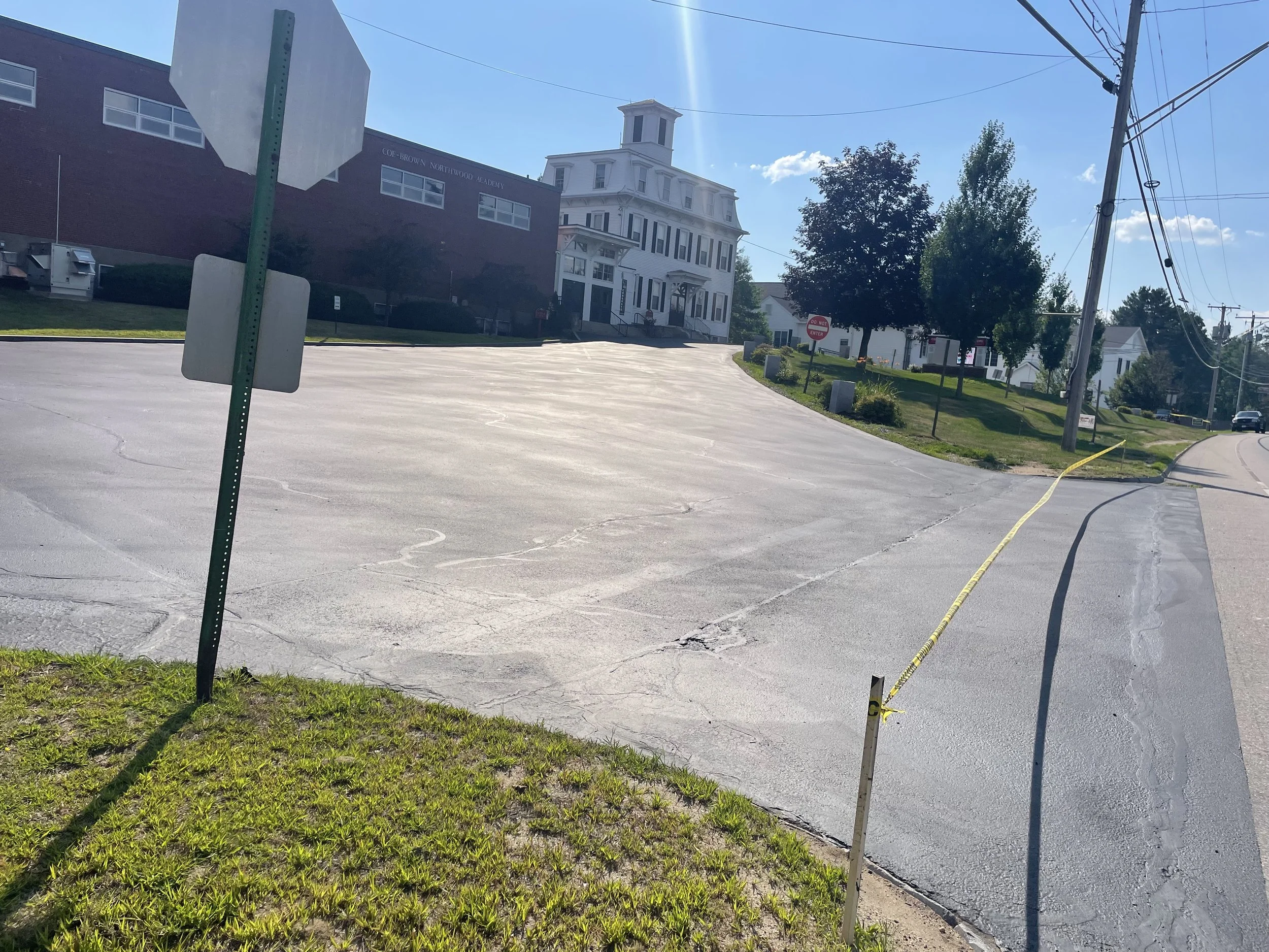 Empty parking lot at the edge of a street, with a yellow caution tape near the sidewalk and a building with a white house and trees in the background.