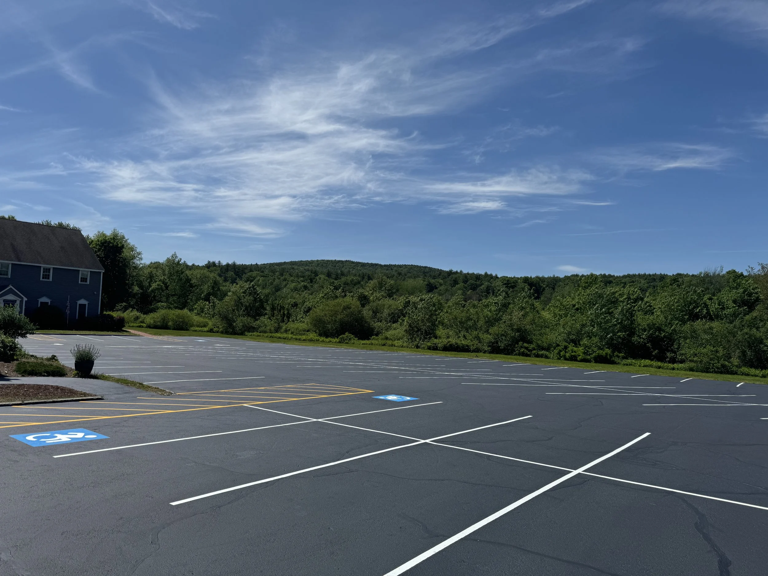 Empty parking lot with marked handicapped spaces on a sunny day, surrounded by greenery and a house on the left.