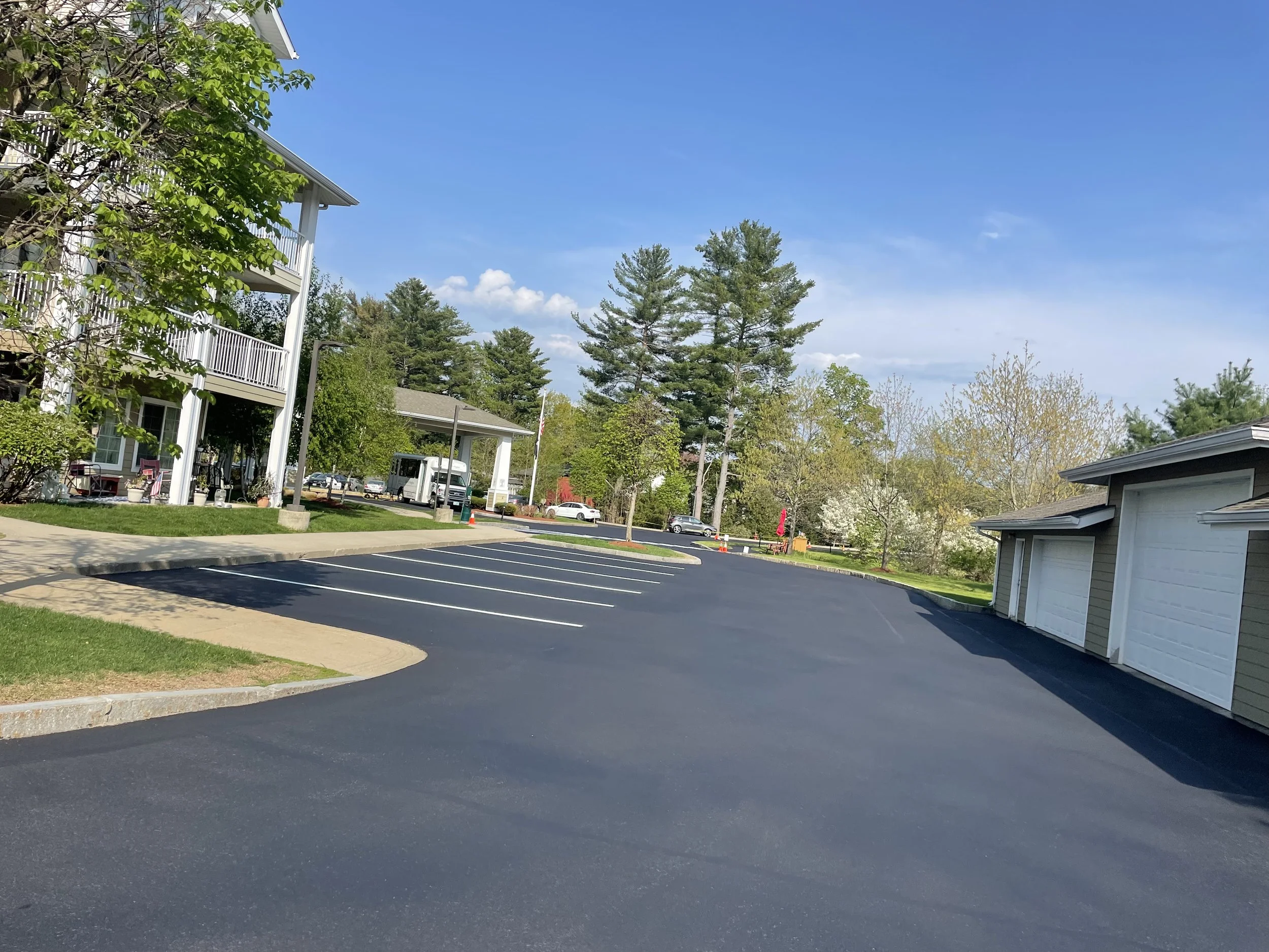 A paved parking lot with marked parking spaces, surrounded by residential buildings and trees with green foliage, under a clear blue sky.