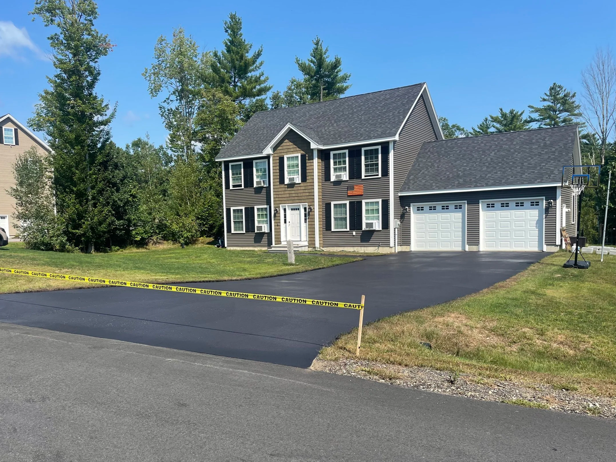 Newly paved driveway in front of a two-story house with black shutters, a basketball hoop to the right, caution tape across the driveway, and green trees in the background.