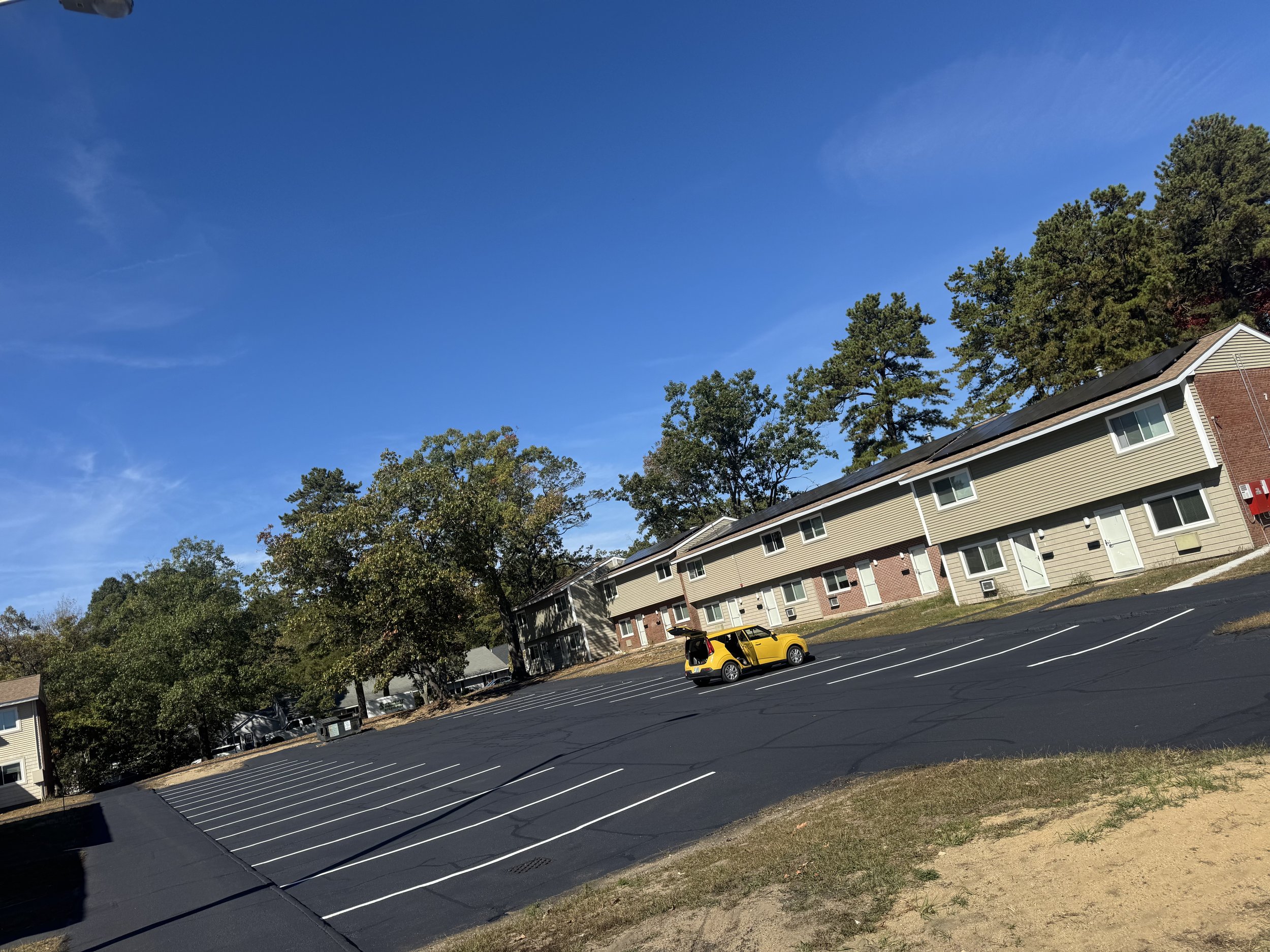 An empty parking lot with a yellow car parked and trees in the background under a bright blue sky.