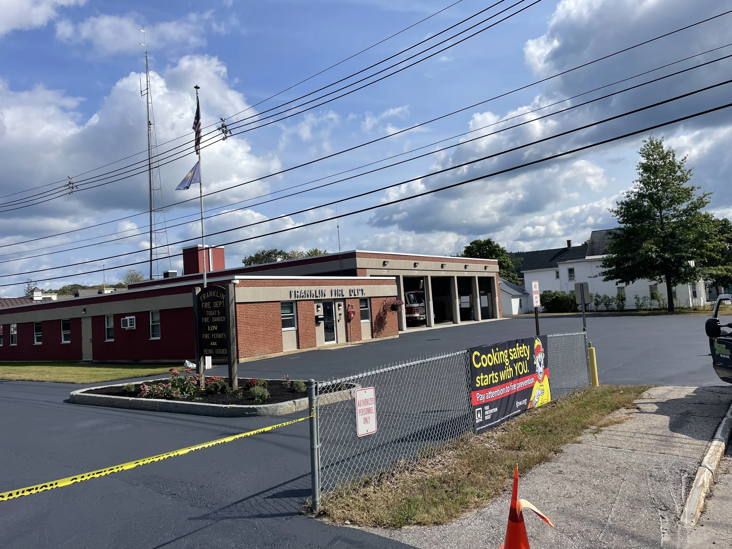 Front view of Franklin Volunteer Fire Department with a parking lot, a chain-link fence, and a banner that reads 'Cooking safety starts with you. Pay attention to fire prevention.' There are power lines in the sky, a flagpole with the American flag and another flag, a small planting bed with flowers, and a single traffic cone.