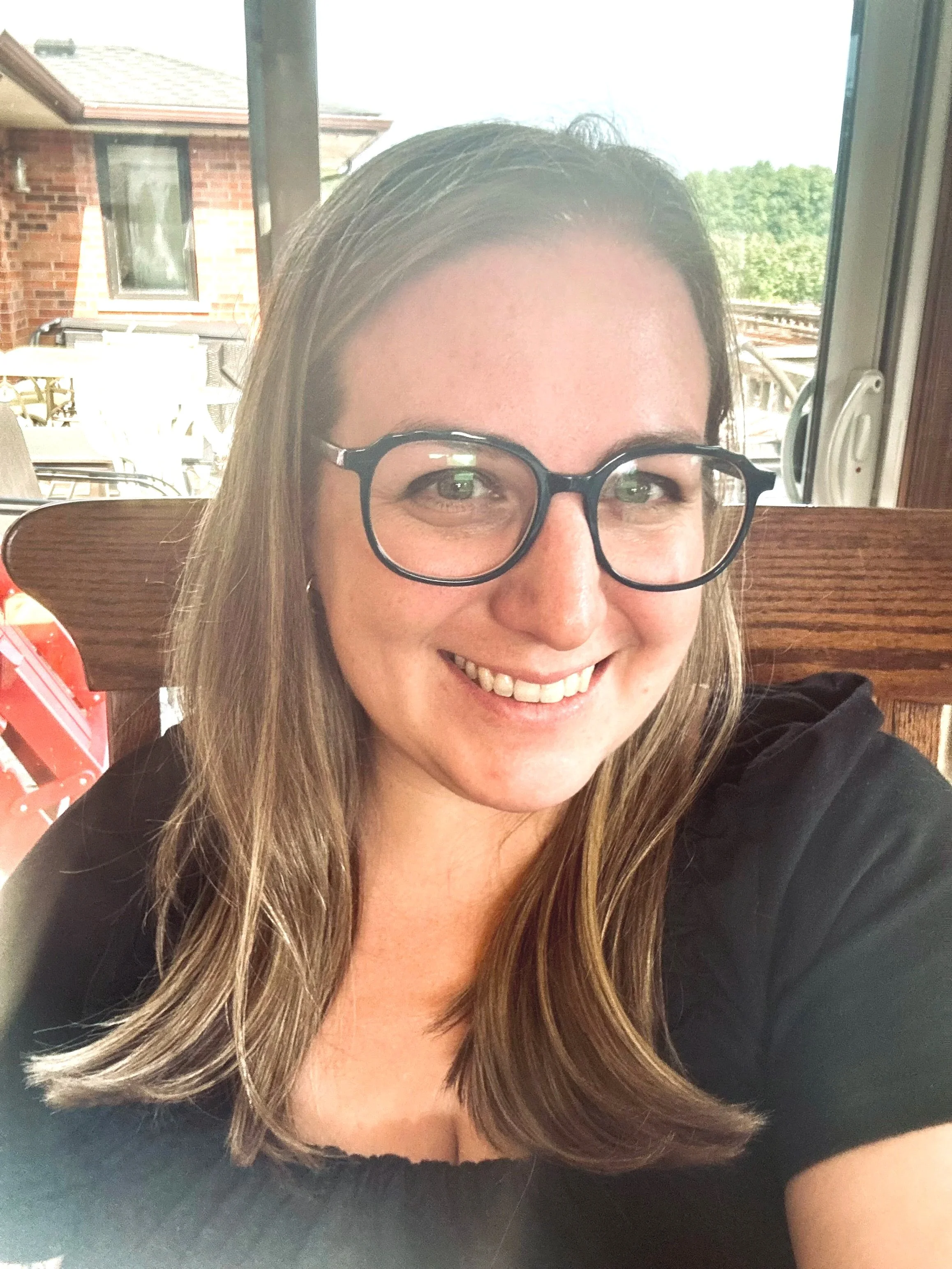 A smiling woman with blonde hair and glasses sitting indoors near a window.