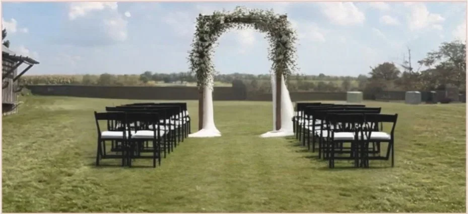 Outdoor wedding ceremony setup with black chairs arranged in rows on either side of a grassy aisle. A decorative arch with greenery and white fabric drapes stands at the end of the aisle under a partly cloudy sky.